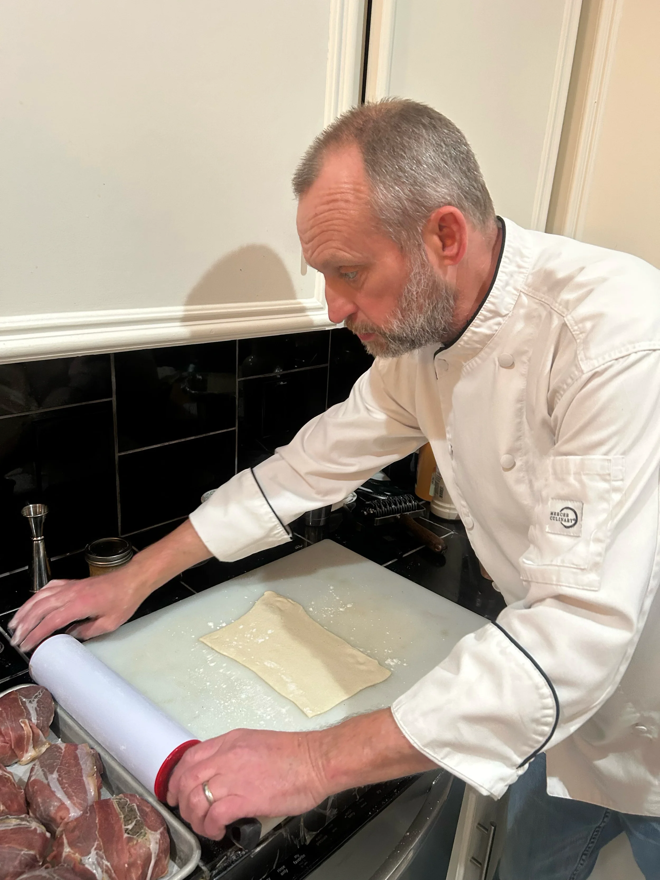 A chef rolling out dough on a countertop with a rolling pin, surrounded by pieces of raw meat.