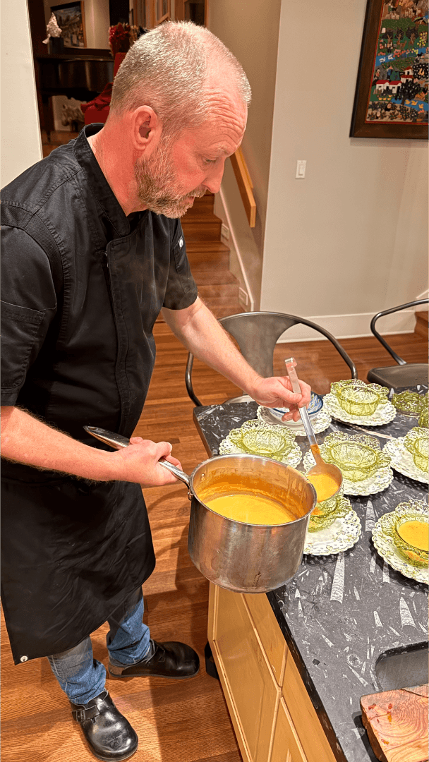 A man in a black apron and jeans serving soup from a pot into bowls on a kitchen counter.
