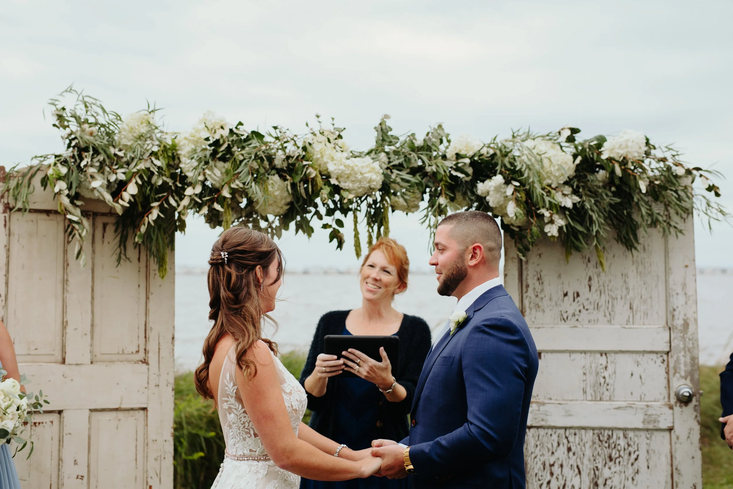 A bride and groom exchanging vows during their outdoor wedding ceremony under a floral arch with white flowers and greenery, with a water body in the background.