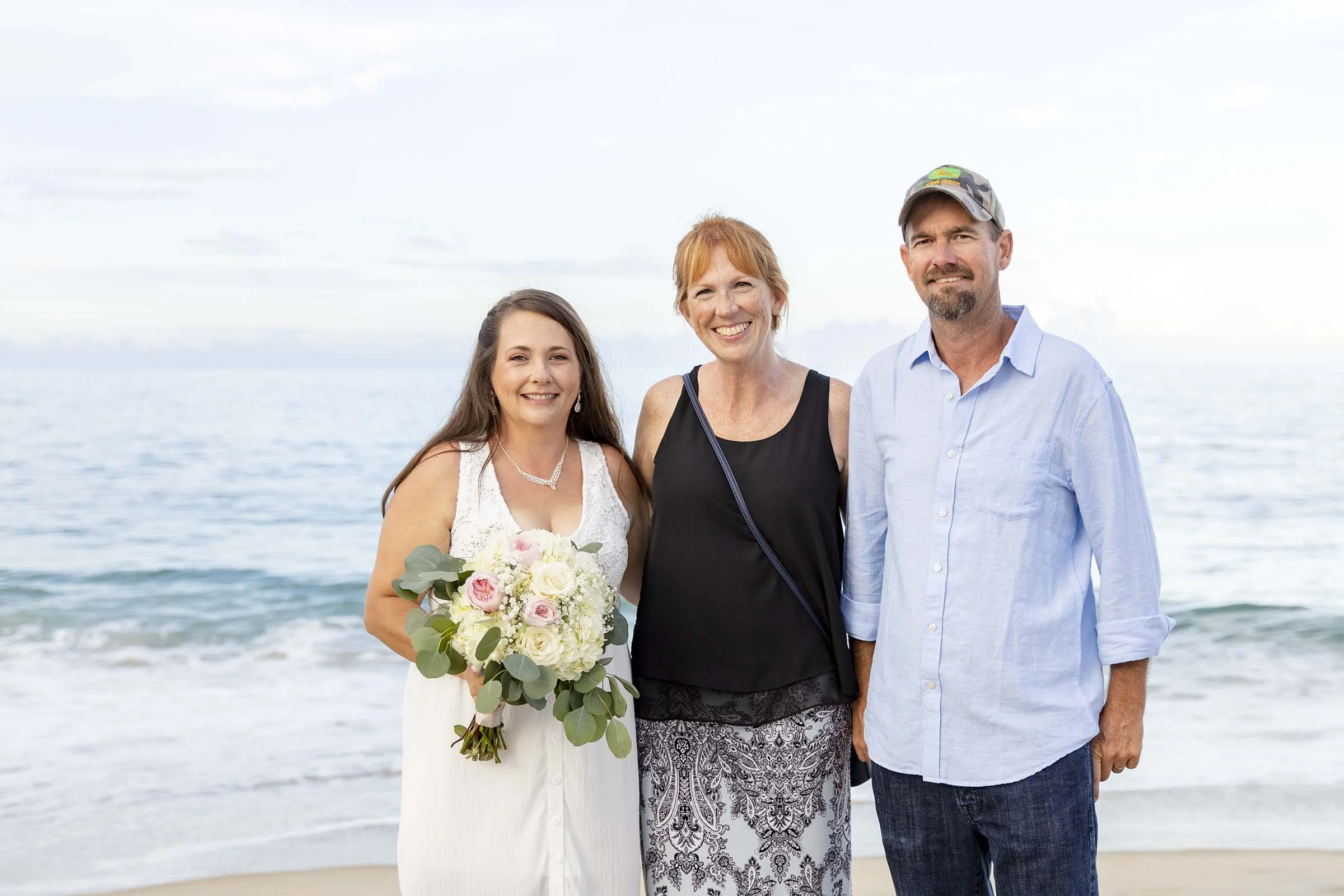 A woman in a white dress holding a bouquet of flowers standing next to a woman in a black top and a man in a light blue shirt, at the beach with the ocean in the background.