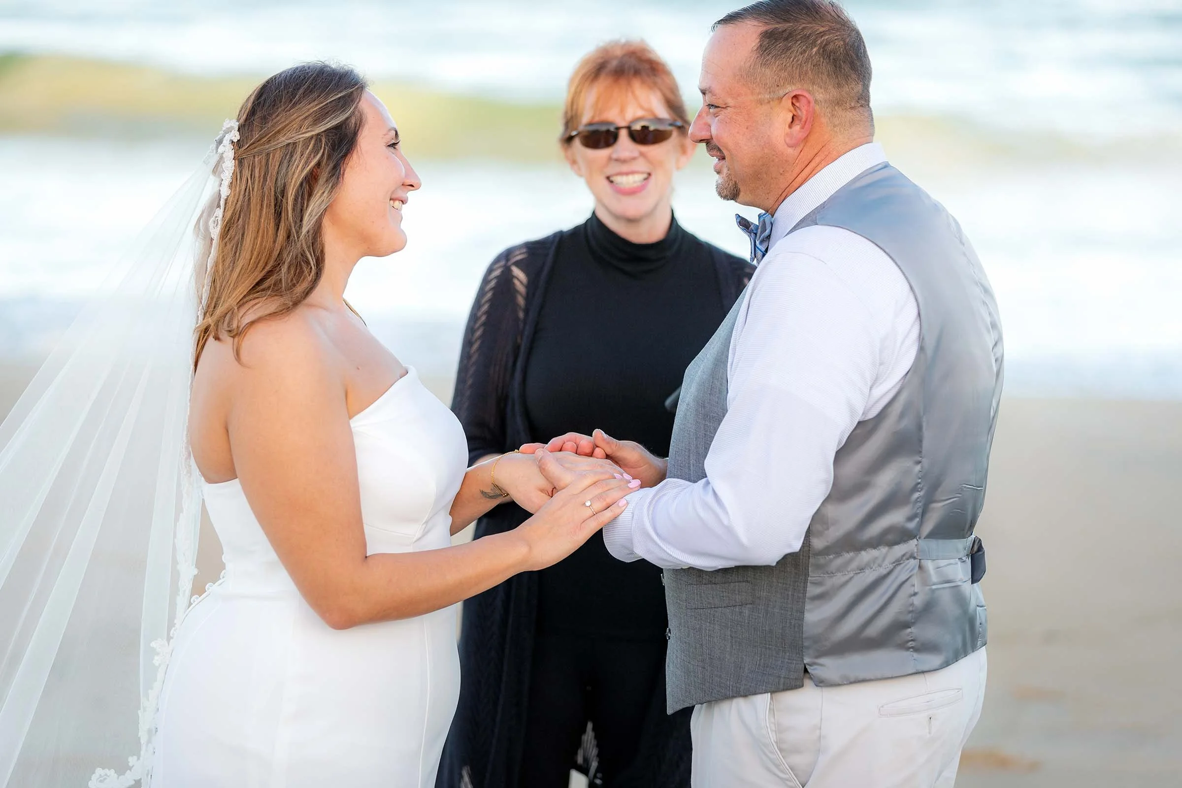 A bride and groom exchange vows on a beach, holding hands and smiling at each other with an officiant standing behind them.