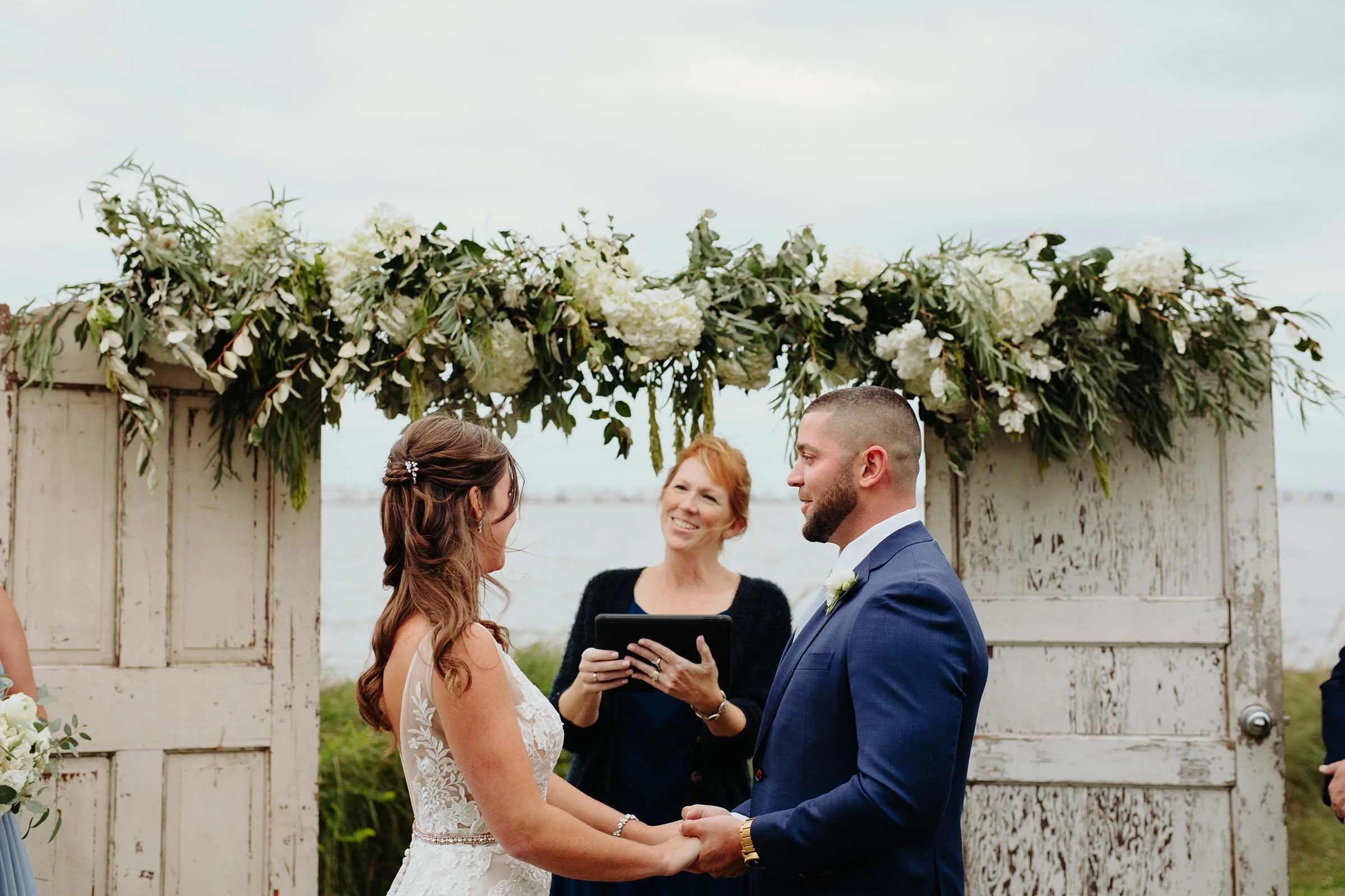 Bride and groom holding hands during wedding ceremony outside, with officiant smiling behind them, in front of a decorated wooden arch with white flowers and greenery.
