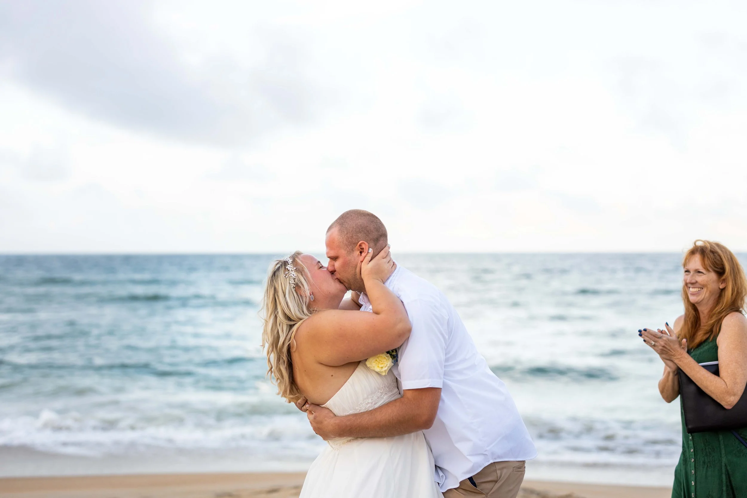 A bride and groom kissing on the beach during their wedding, as OBX wedding officiant claps and smiles nearby.