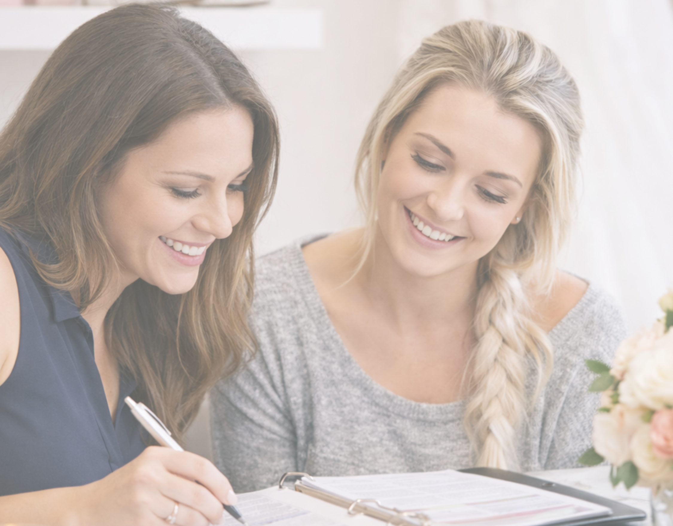 Two women smiling and looking at documents or a planner on a table, one signing with a pen, in a bright, casual setting.