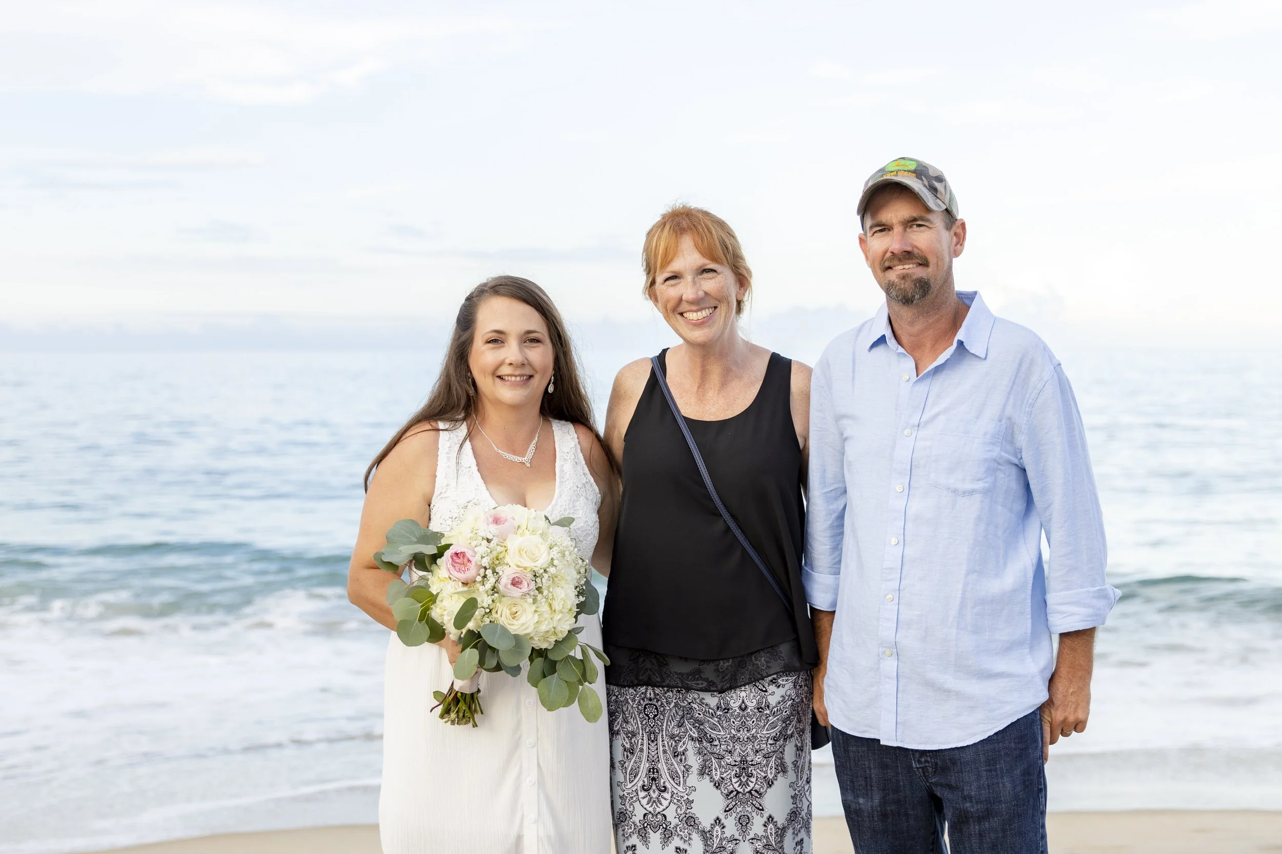 Three smiling people standing on the beach near the ocean, with a woman holding a bouquet of flowers.