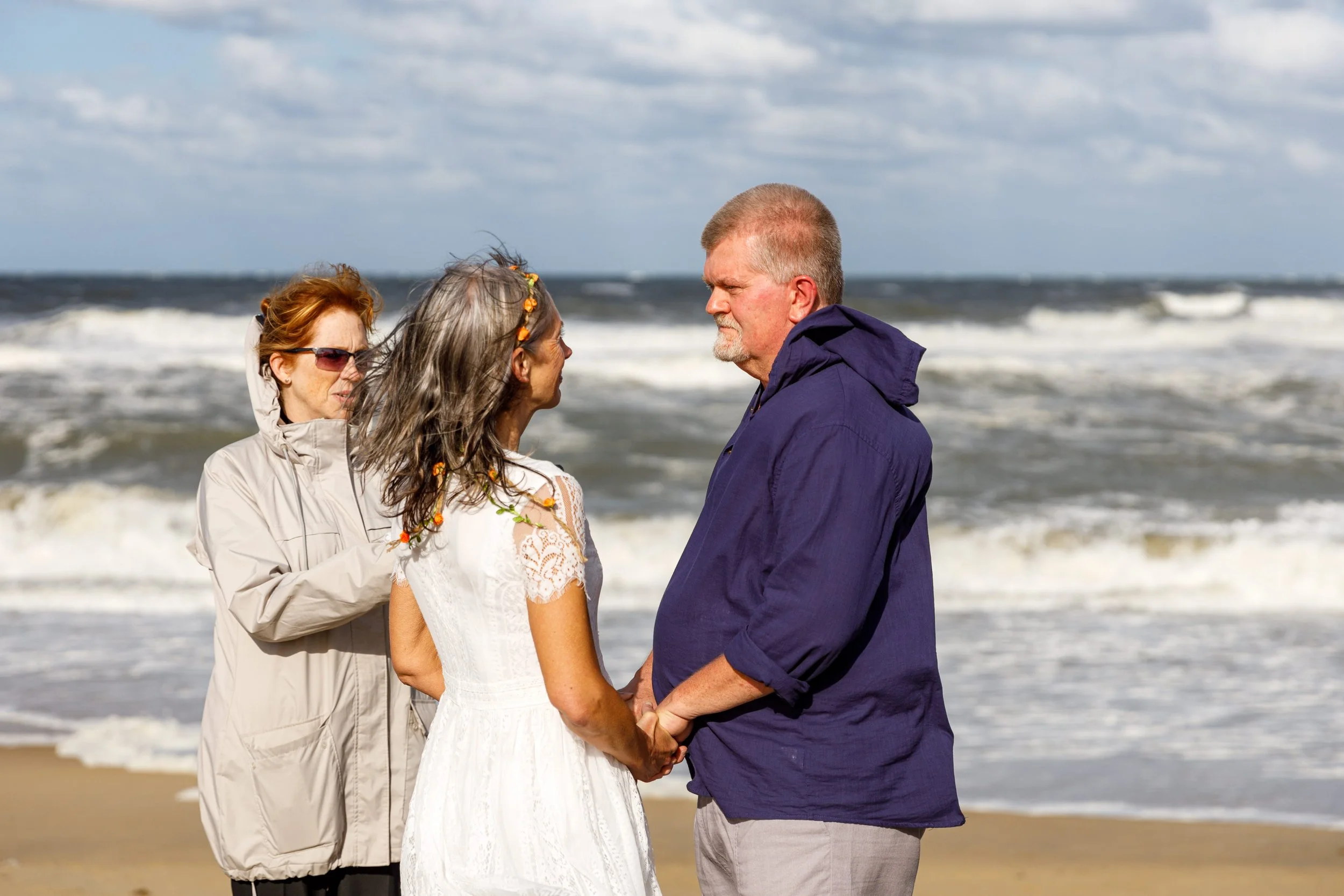 A couple holding hands on the beach facing each other, with a woman officiant standing behind them. The ocean and cloudy sky are in the background.
