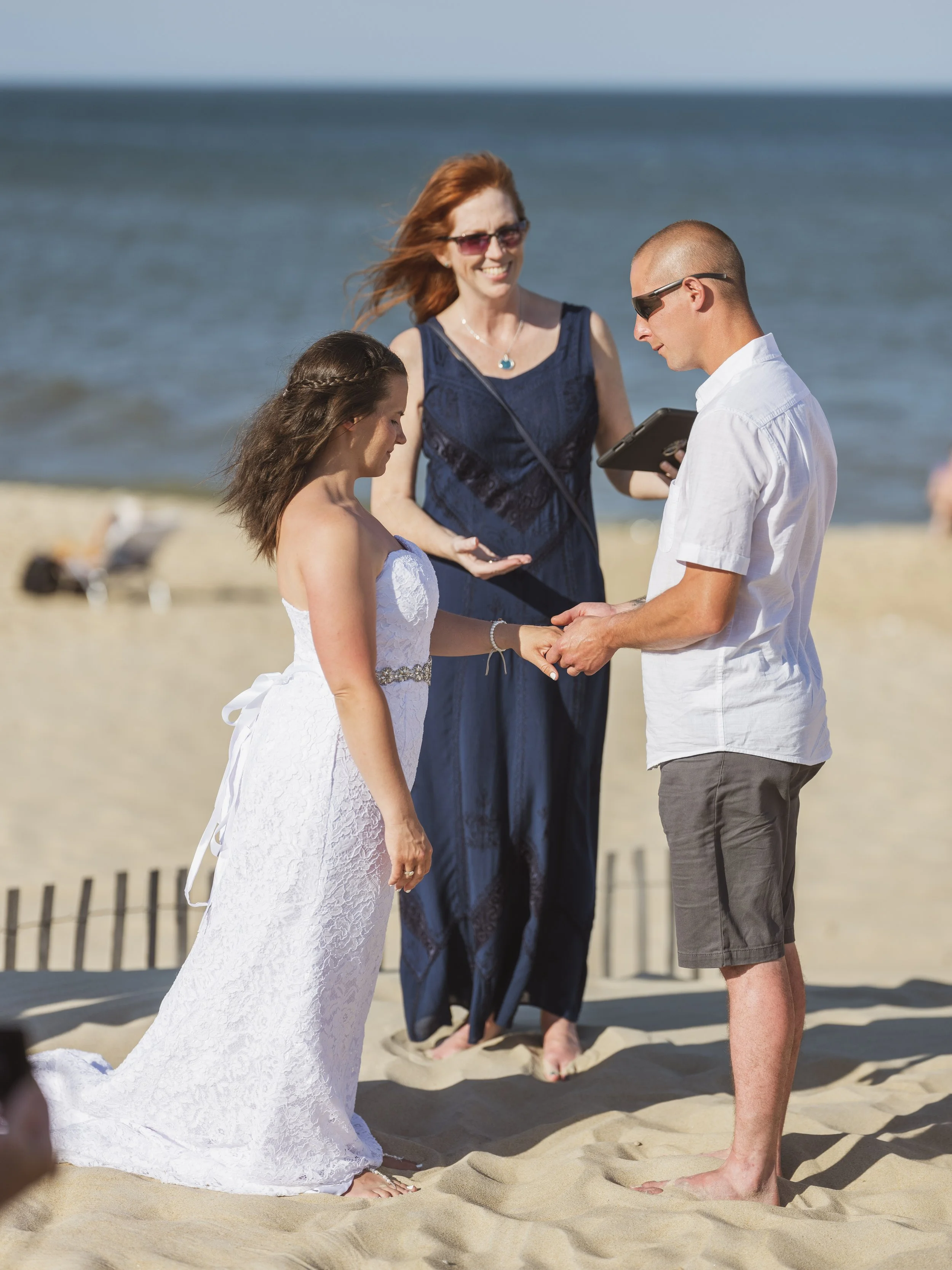 A couple getting married on the beach, standing barefoot in the sand, with a woman officiating at the ceremony. The bride is in a white wedding dress, and the groom is in a white shirt and gray shorts. The ocean is in the background.