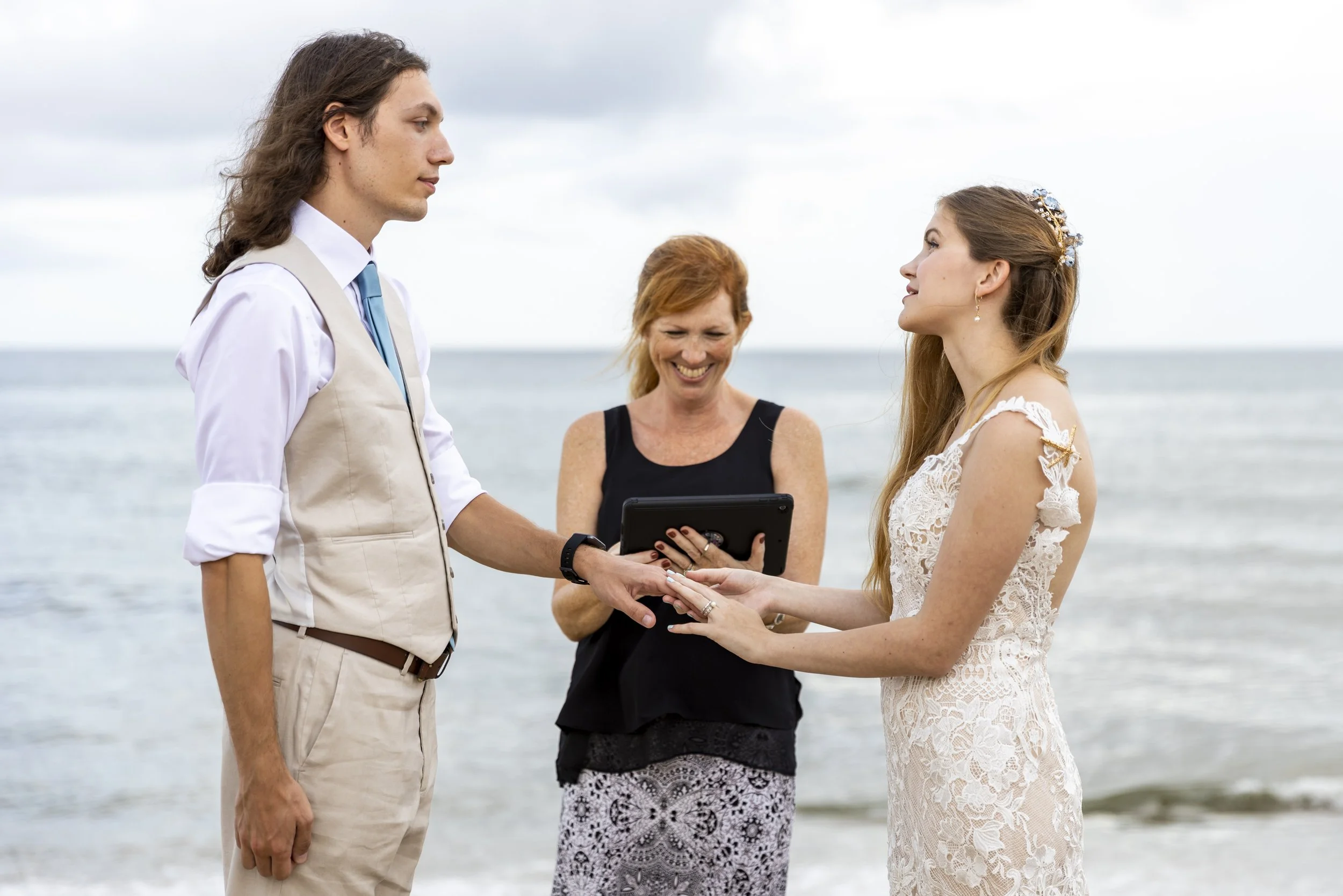 A couple exchanging wedding vows on the beach with an officiant smiling in the background.