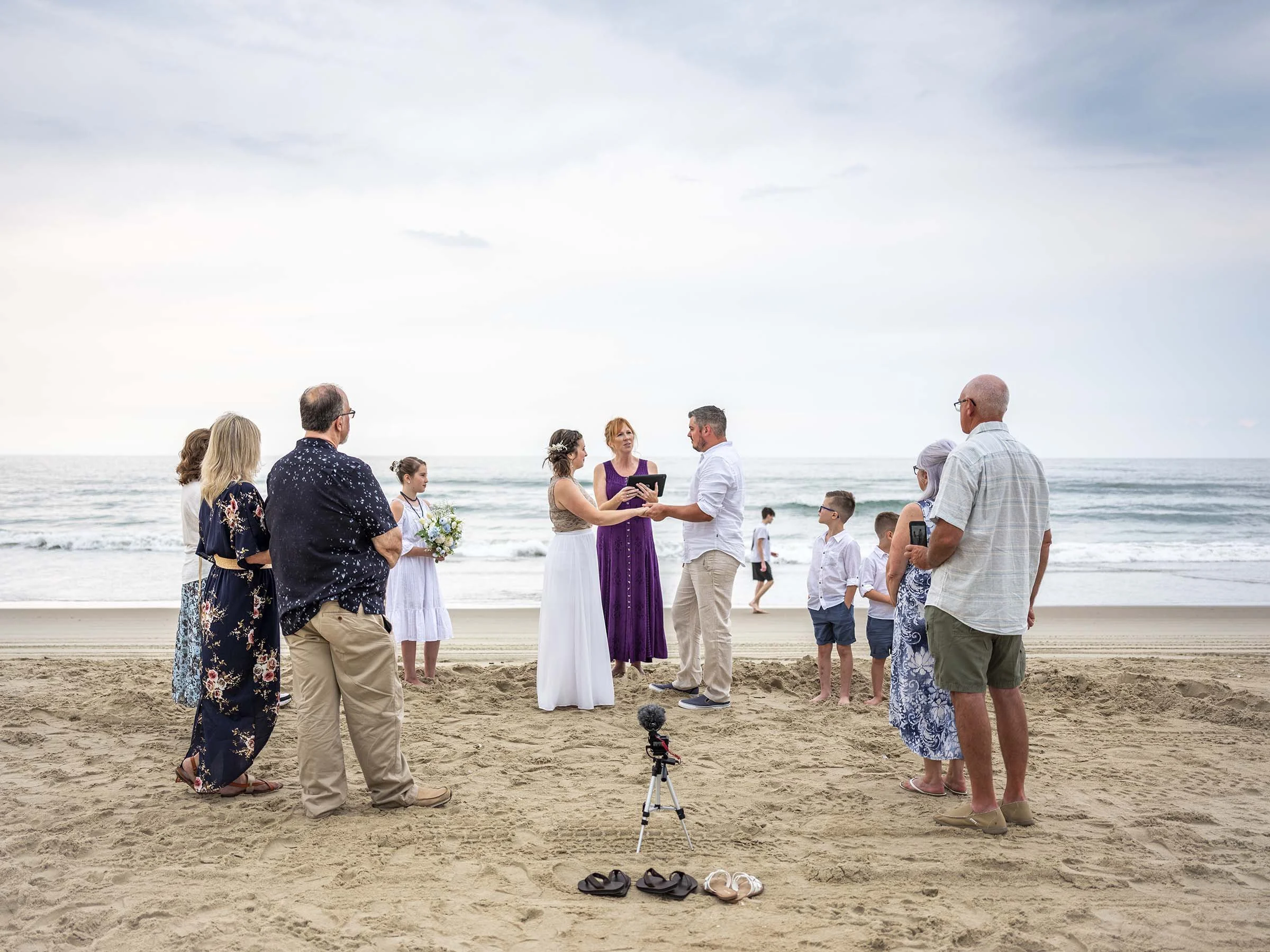 A beach wedding ceremony with a bride, groom, officiant, and guests standing in a circle near the ocean under cloudy sky.