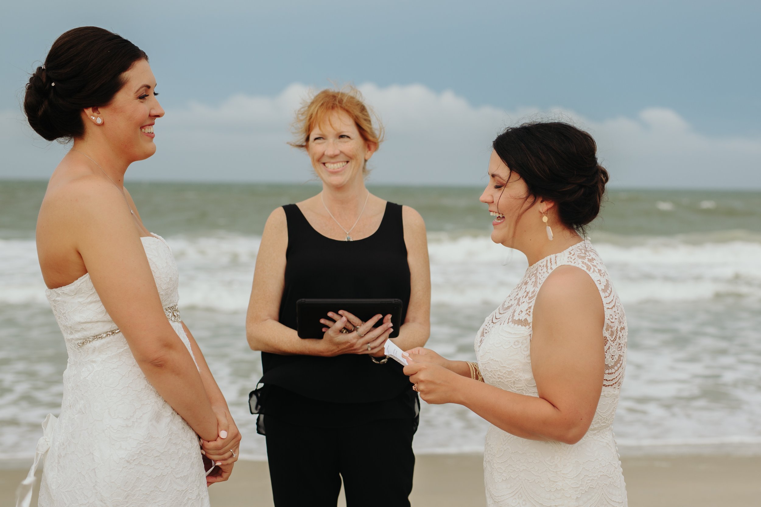 Two women in wedding dresses smiling at each other on the beach during a wedding ceremony with the officiant standing between them, holding a small piece of paper, ocean waves in the background.