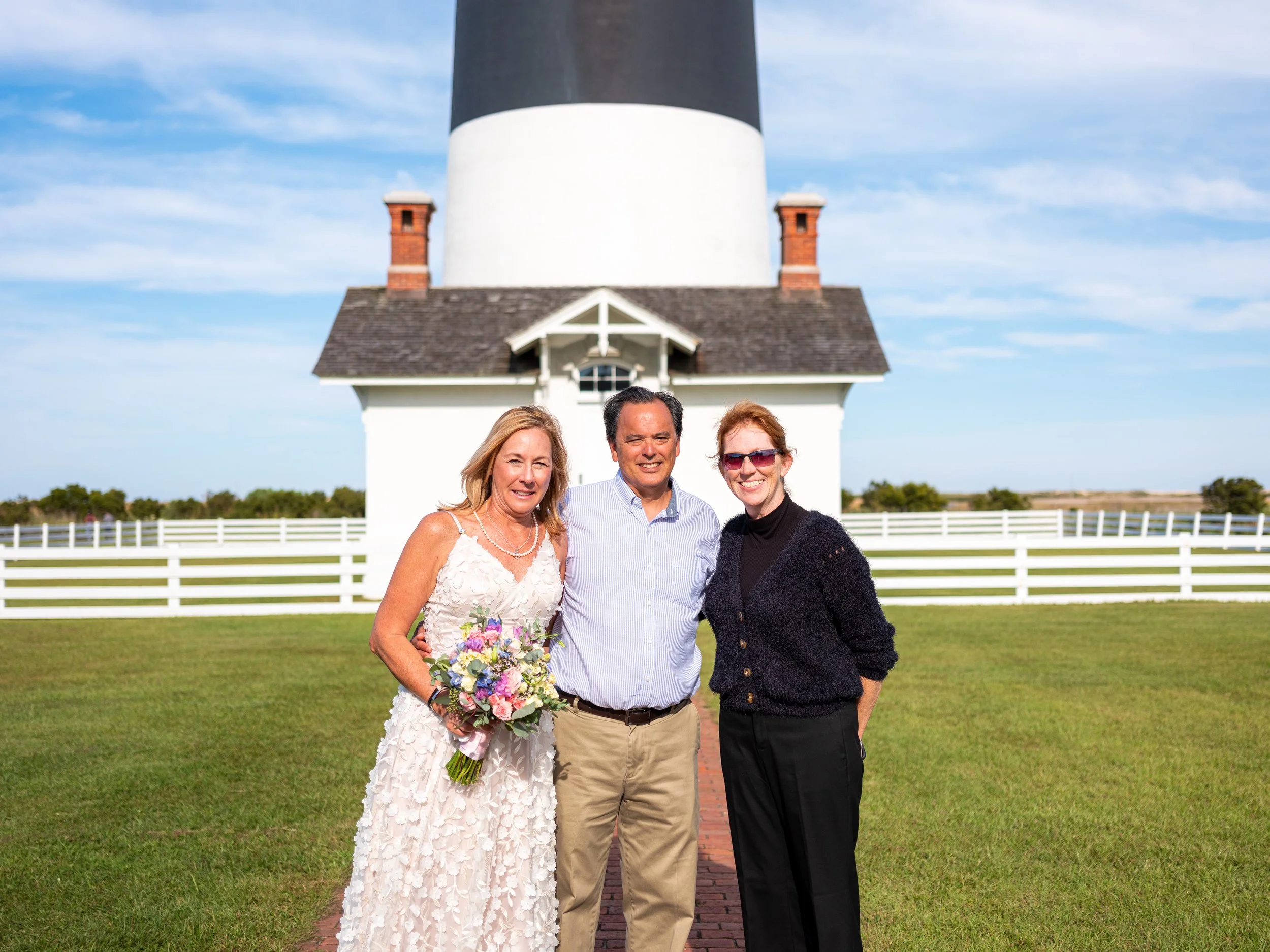 Three people standing on a brick path in front of a white building with a black and white lighthouse structure, a grassy field, and a mostly blue sky with some clouds in the background. The woman on the left is holding a bouquet of flowers and is wea