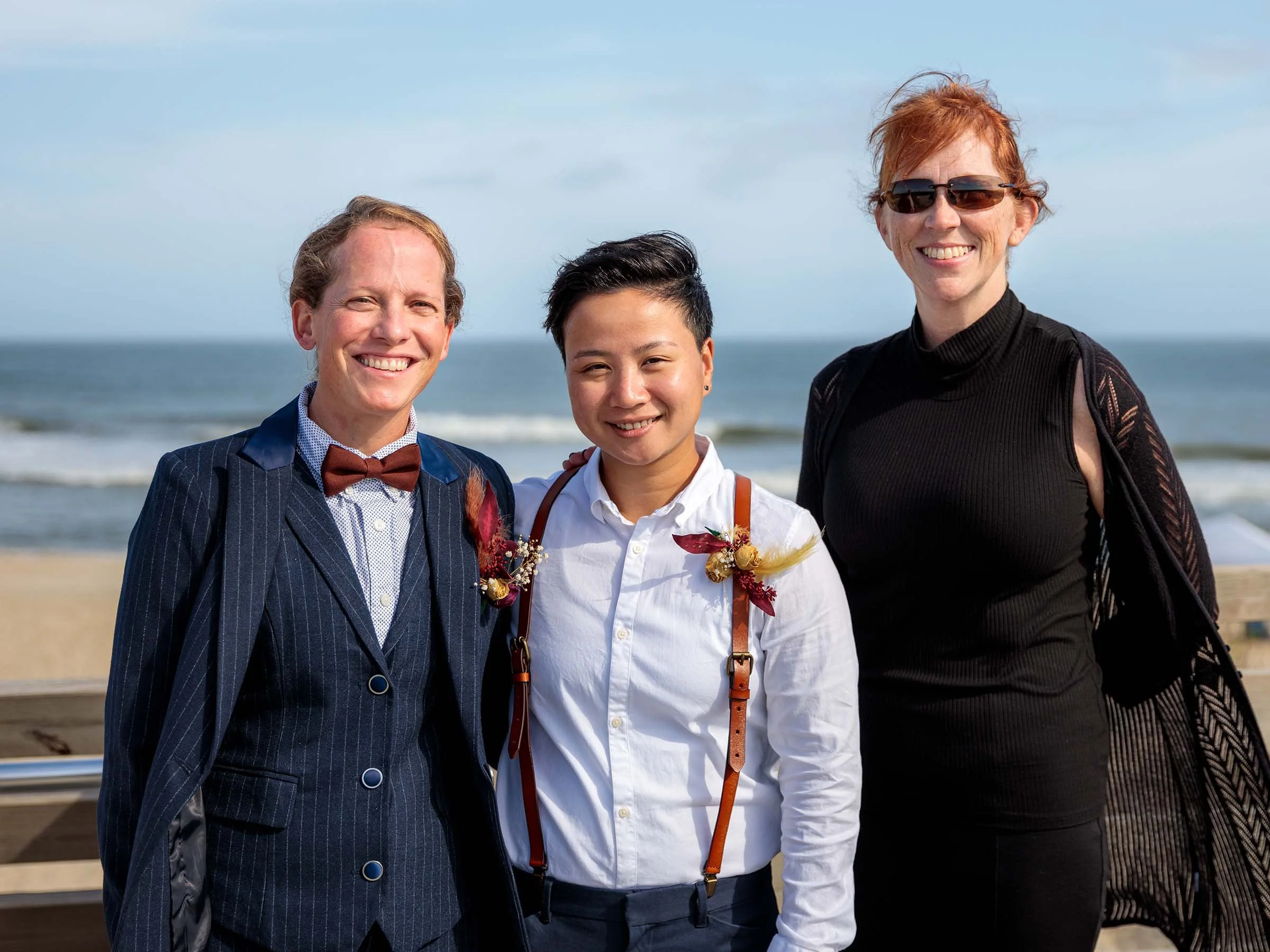 Three smiling people standing on the beach near the ocean, dressed in a mix of formal and casual attire.