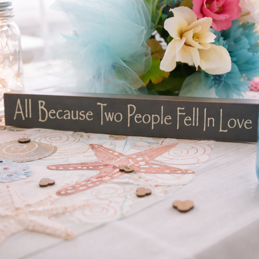A decorative sign reading 'All Because Two People Fell In Love' on a table with a starfish design, surrounded by flowers and small wooden hearts.