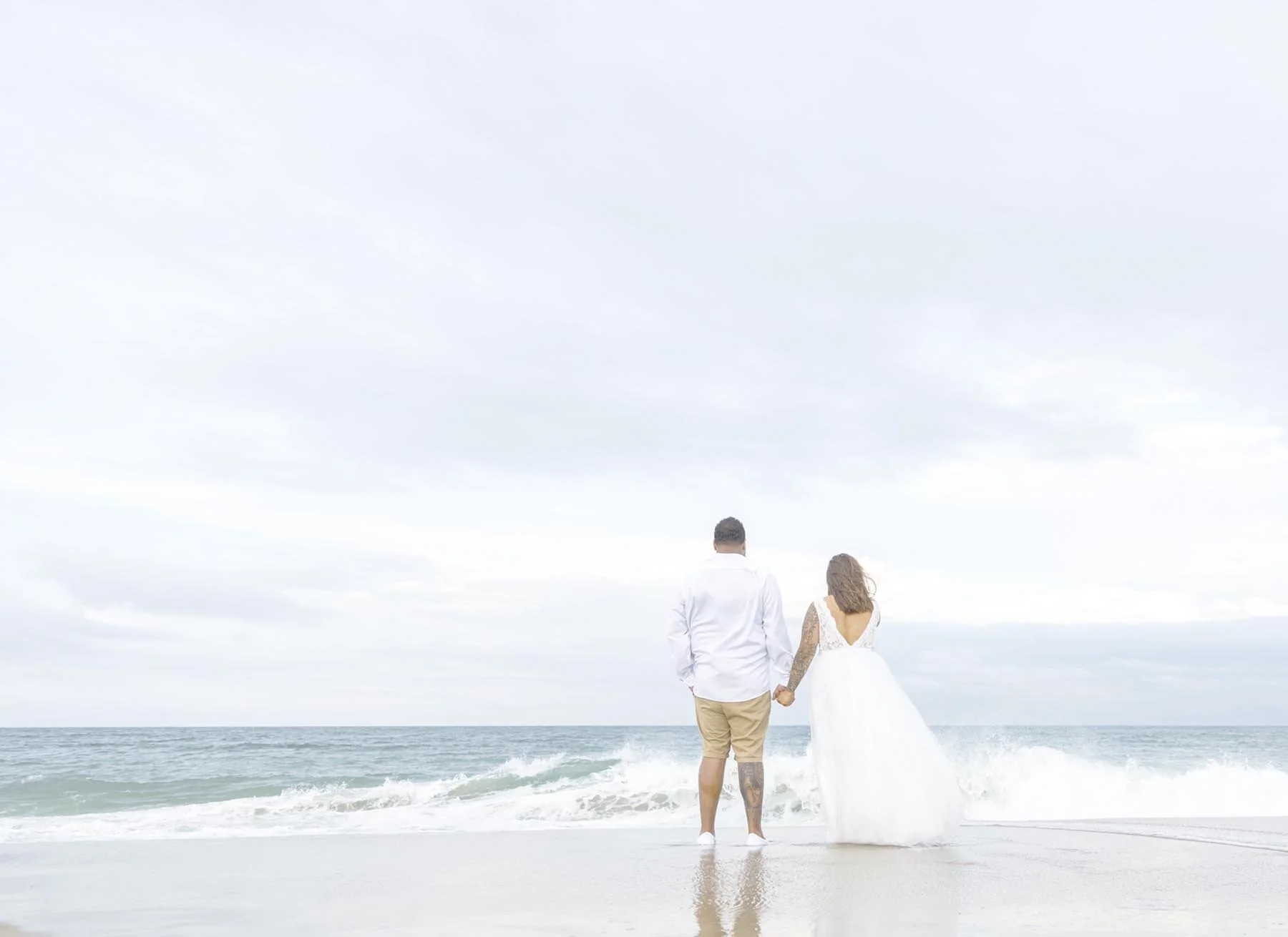 a bride and groom holding hands, standing by the OBX surf.