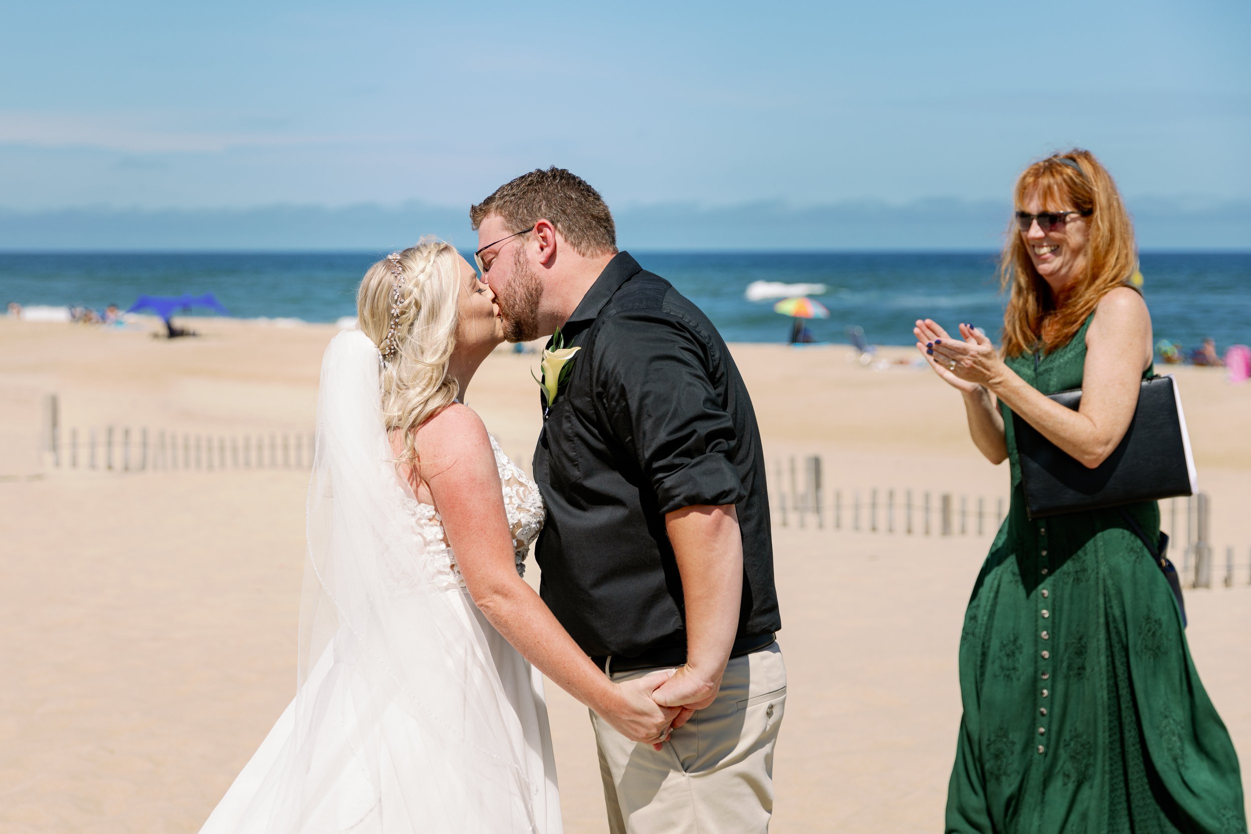A bride and groom kiss on a beach with an officiant clapping in the background.