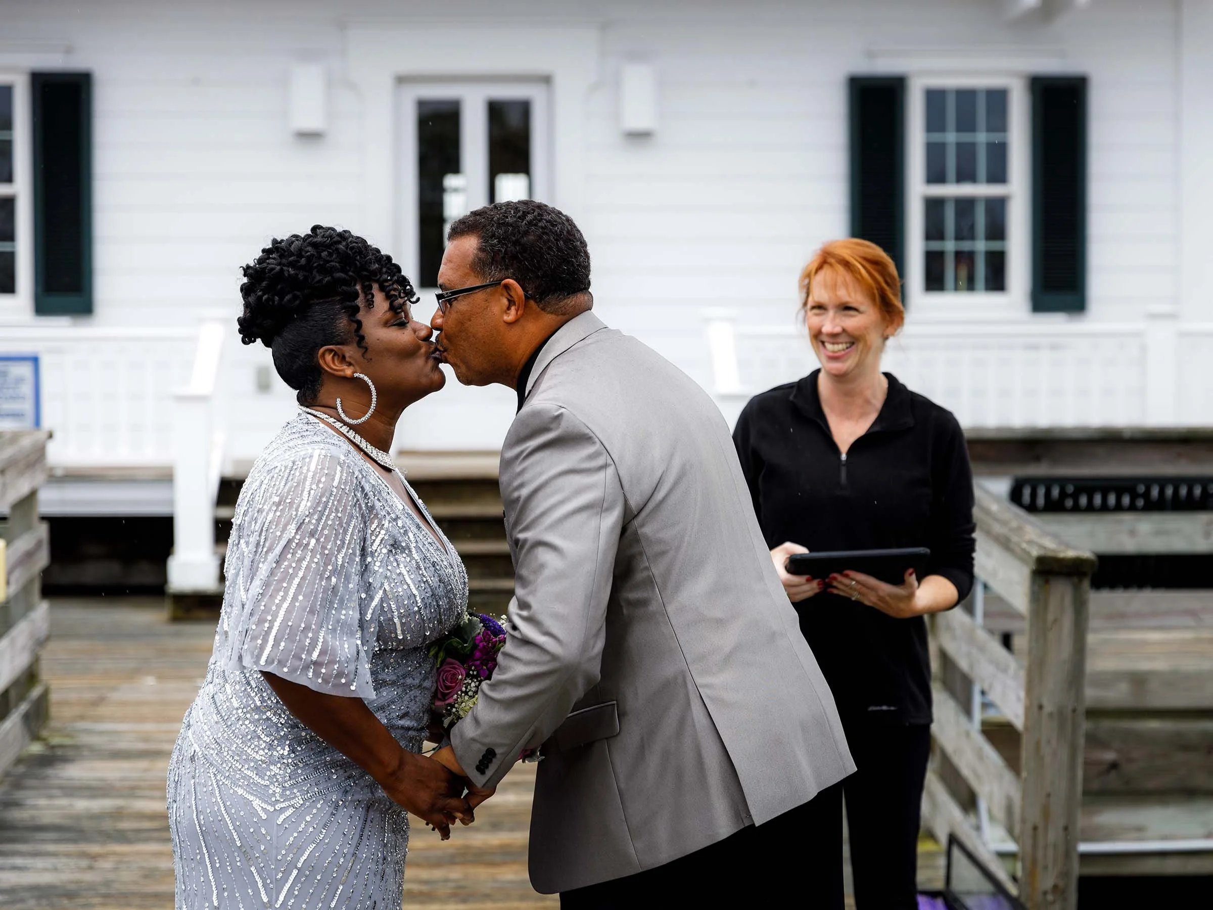 A same-sex couple, a woman and a man, are holding hands and kissing outdoors in front of a white house, while a woman witnesses with a smile.