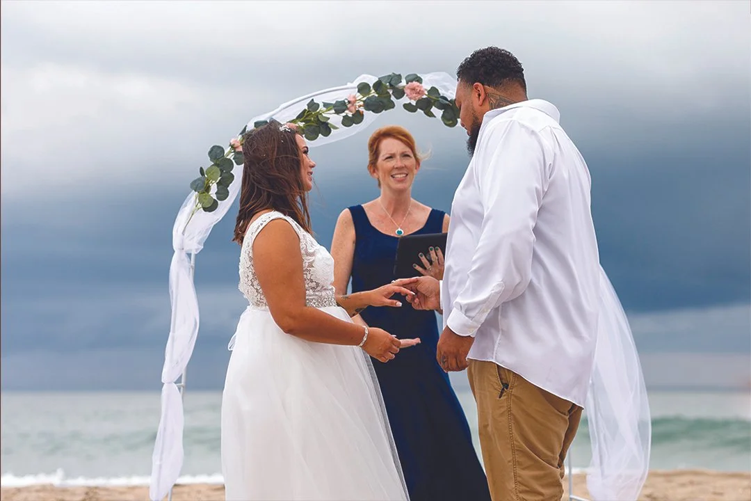 A couple gets married on the beach under a floral arch, with a woman officiating the ceremony, as dark clouds gather in the sky.