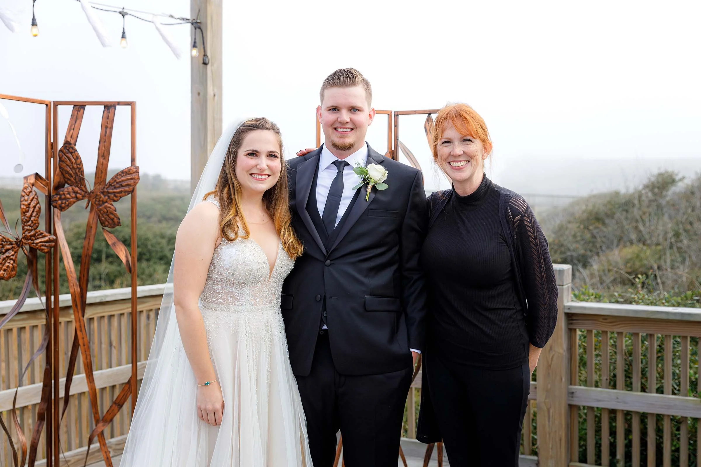 Bride, groom, and woman smiling outdoors on a wooden deck with decorative butterflies and string lights.