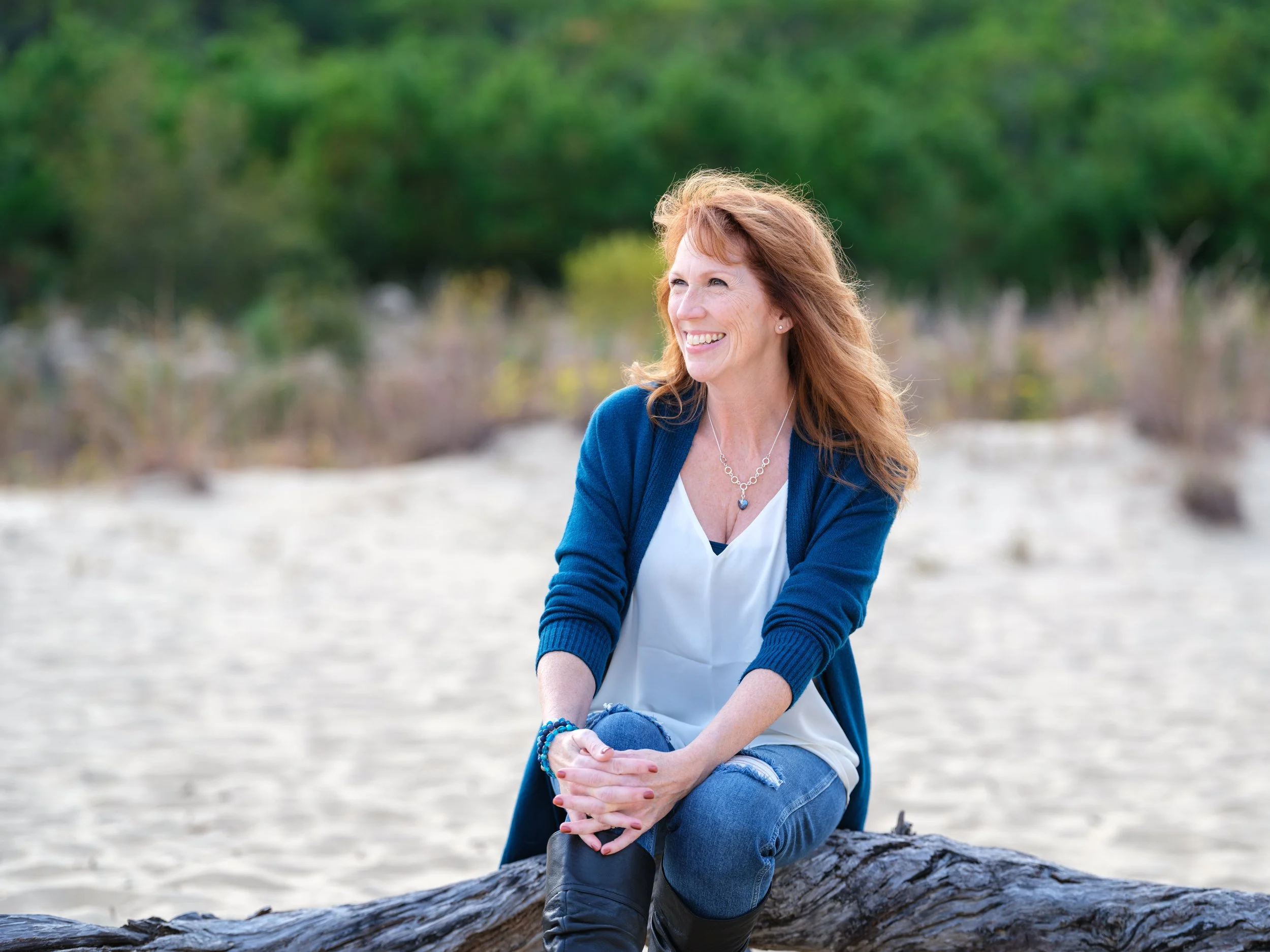 A woman with red hair, wearing a blue cardigan, white blouse, and jeans, sitting on a log on a sandy beach, smiling and looking to the side, with green trees in the background.