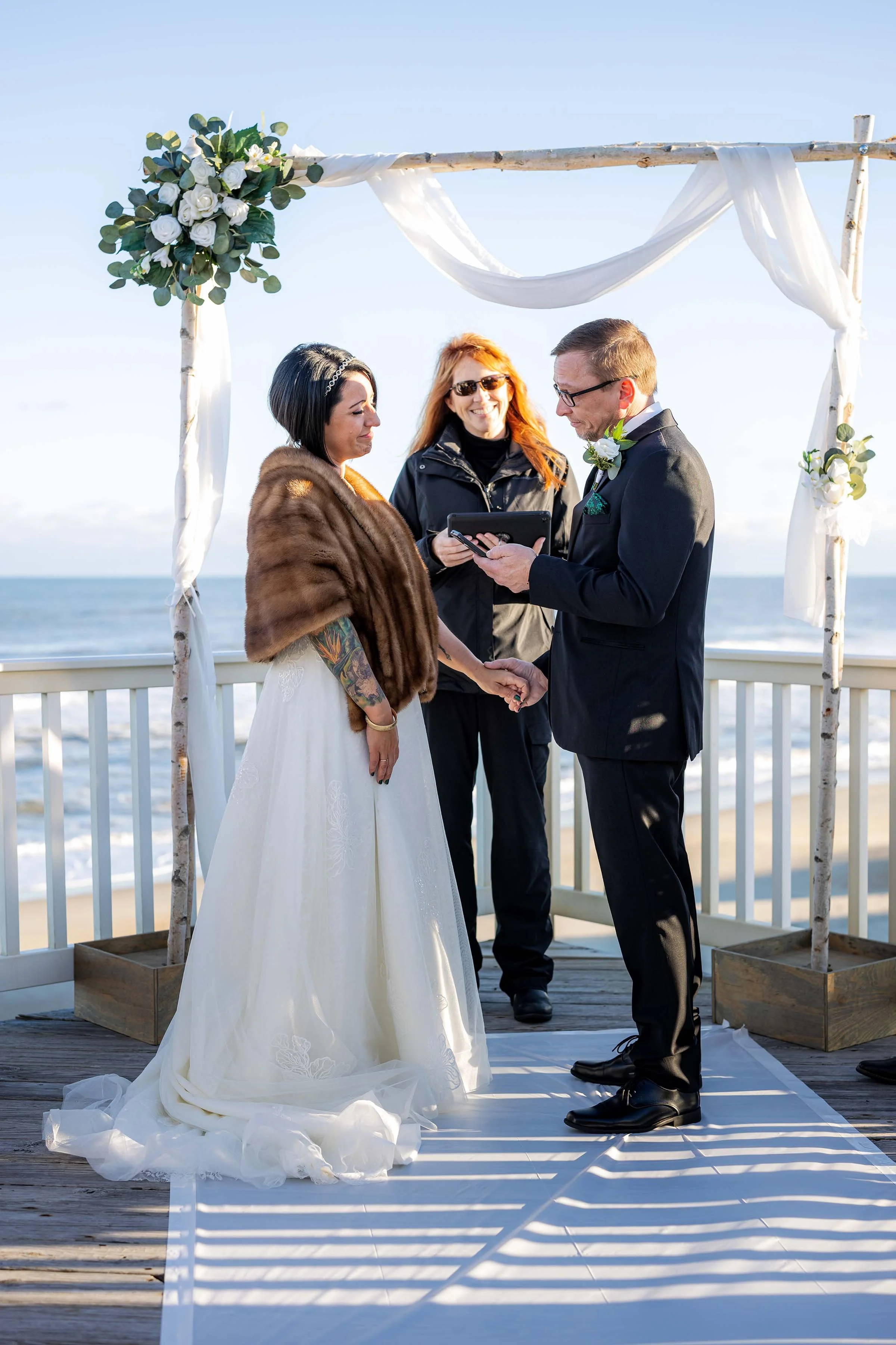 A wedding ceremony taking place on a beach with an officiant holding a tablet, a bride and groom holding hands under a wooden arch decorated with white fabric and flowers, ocean in the background, bright sunlight.