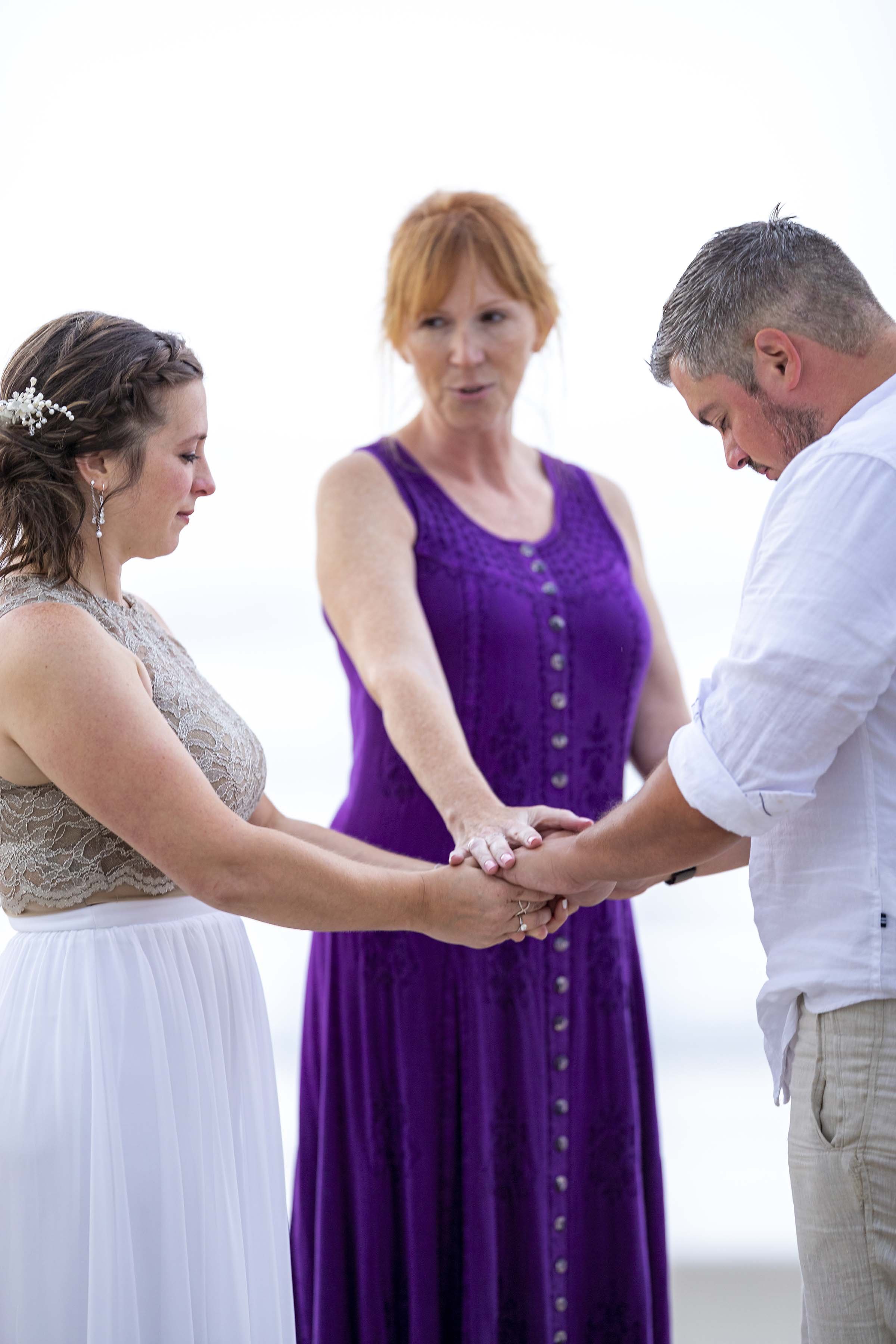 A couple holding hands during a wedding ceremony, with an officiant in the background.