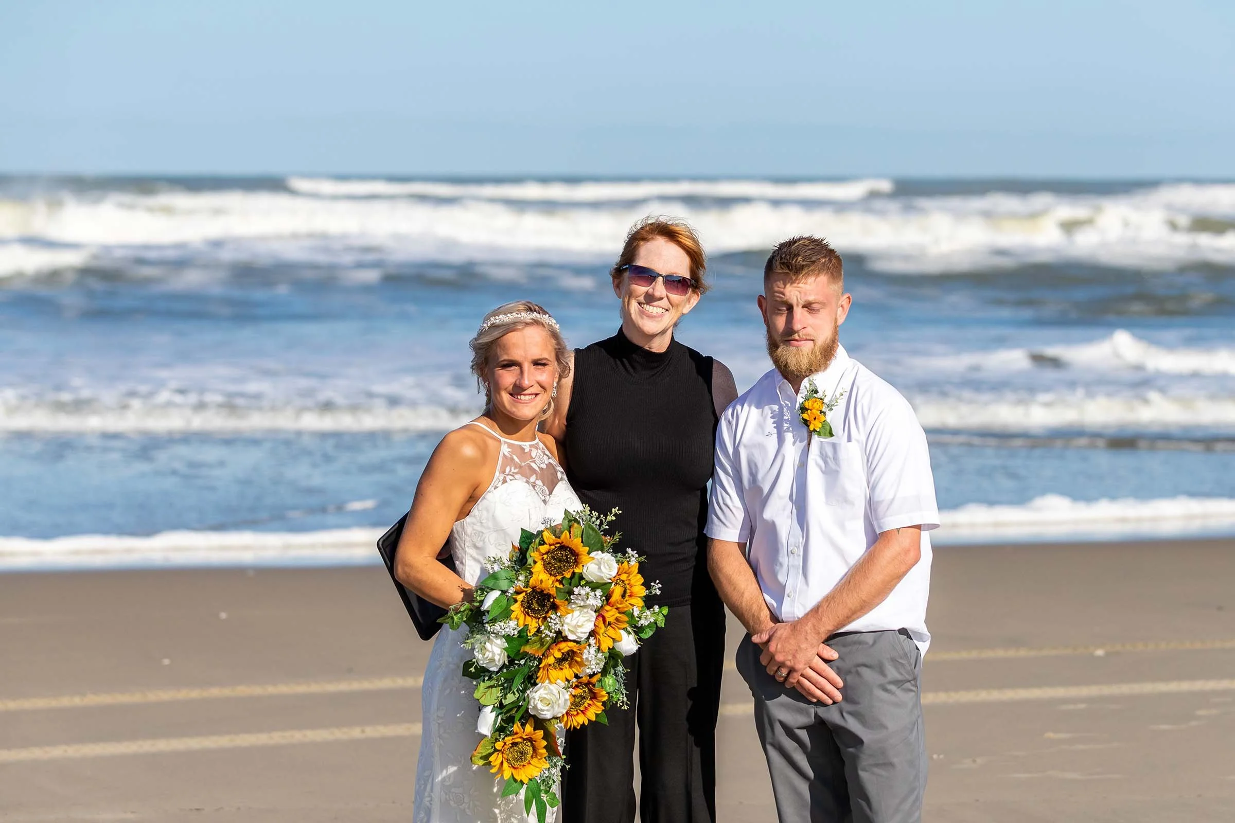 A bride, a woman, and a groom, a man, standing on a beach with ocean waves in the background. The bride is wearing a white wedding dress and holding a sunflower bouquet. The woman in the middle is wearing sunglasses and a black outfit. The man is wea