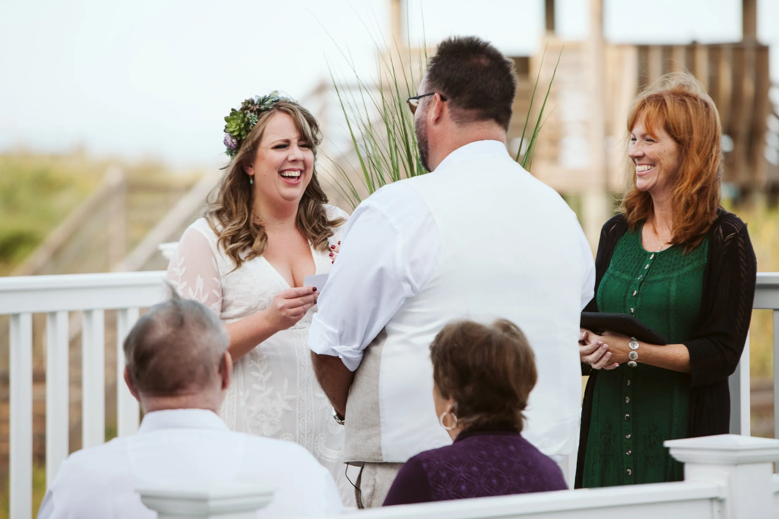 A woman in a white dress with a flower crown reads vows during a wedding ceremony outdoors, with a man facing her, and two women listening and smiling in the background.