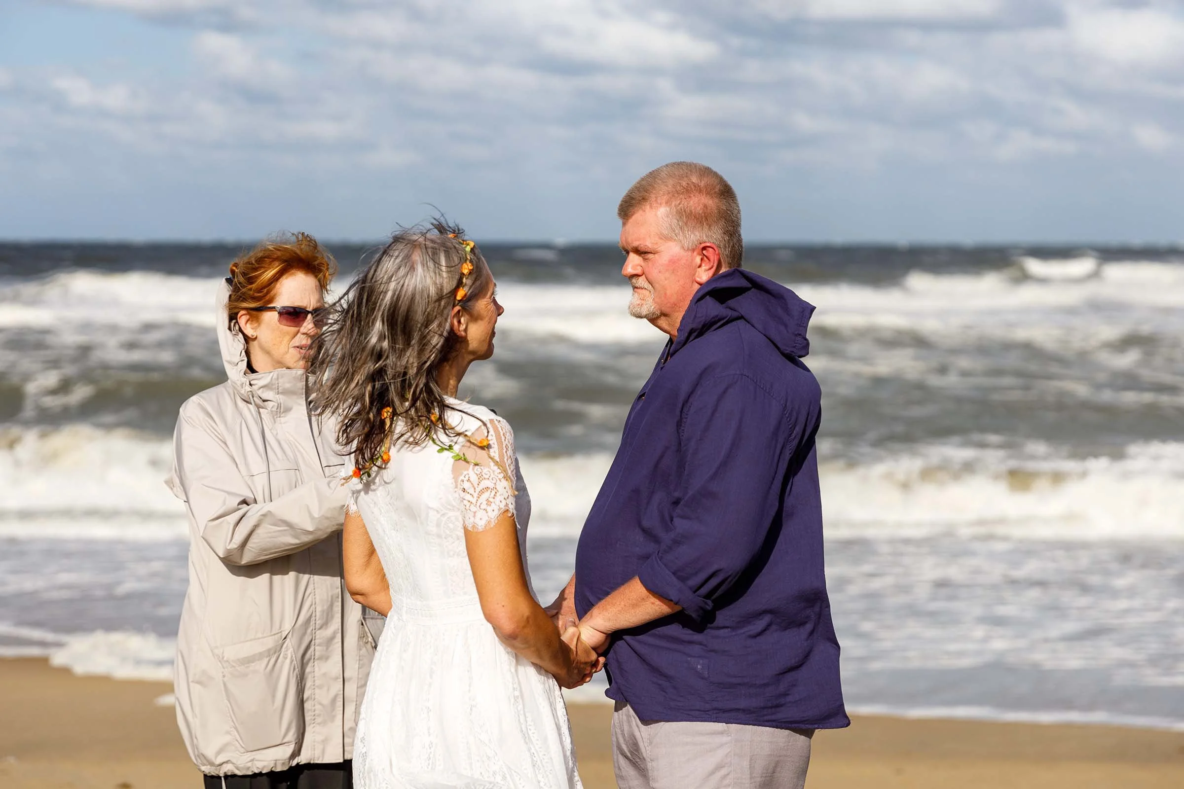 A wedding ceremony taking place on a beach with three people standing face to face, holding hands, with ocean waves in the background. One woman in a white dress and floral headband, a man in a dark blue jacket, and another woman in a light jacket.