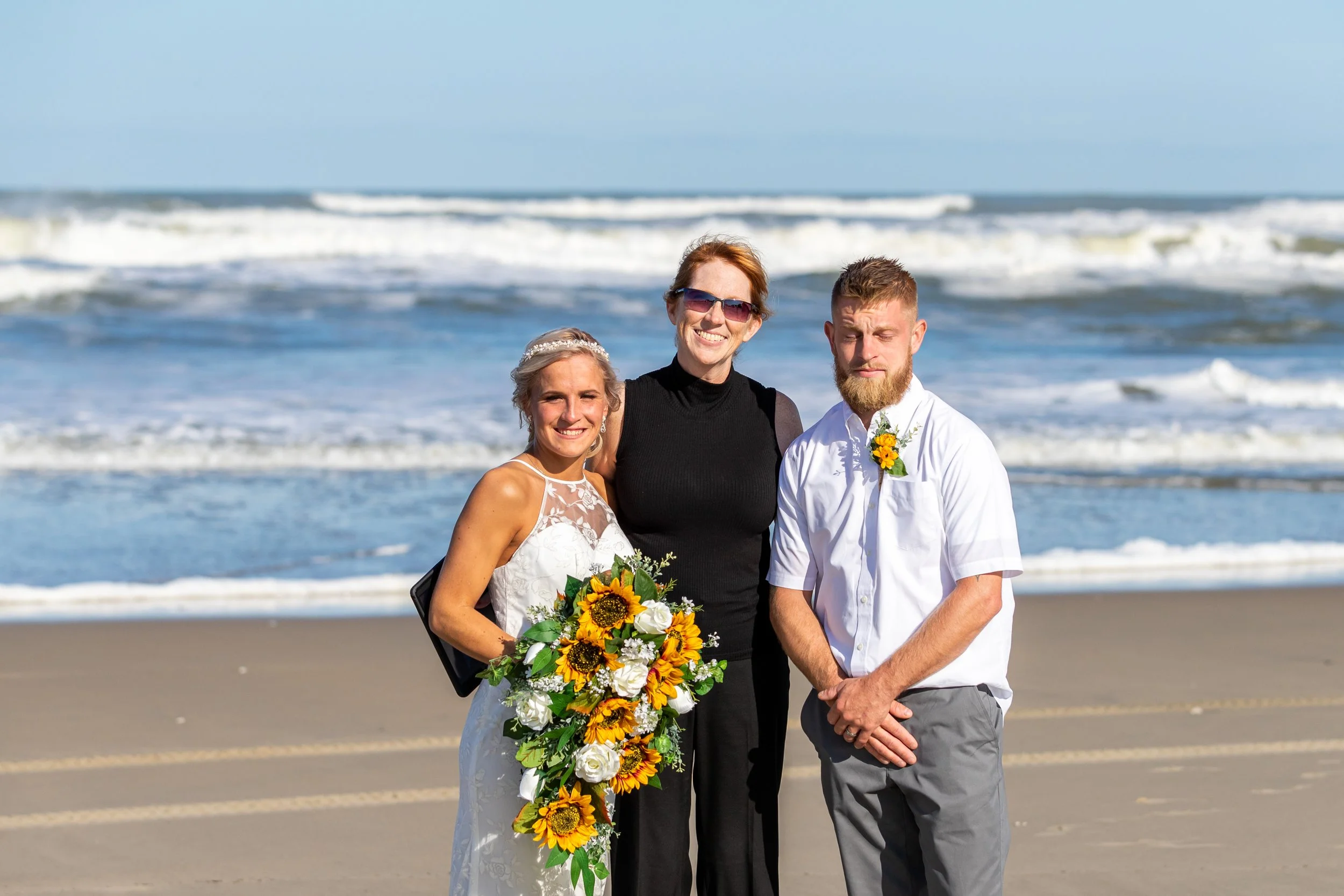 Three people standing on a sandy beach with ocean waves in the background, dressed for a wedding, smiling.
