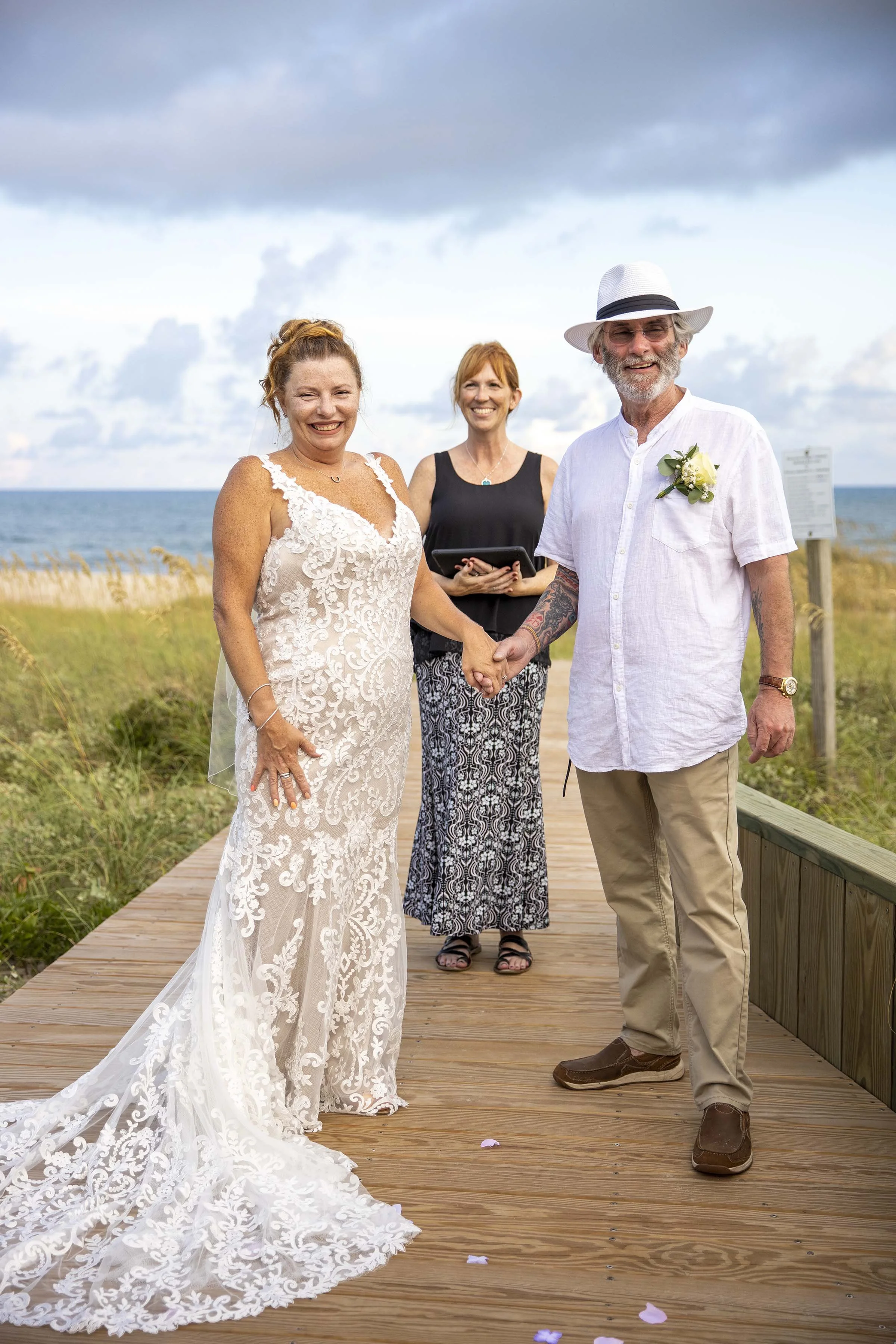 A couple is getting married outdoors on a wooden boardwalk near the beach, holding hands and smiling. The bride wears a white lace wedding gown, and the groom wears a white shirt, beige pants, and a white hat. An officiant stands behind them, holding