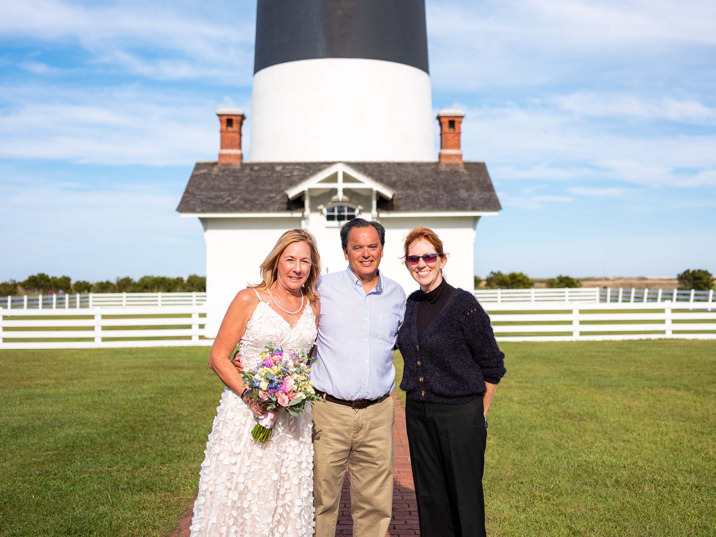 Three people standing arm-in-arm outdoors in front of a white house with a black roof and a lighthouse tower.