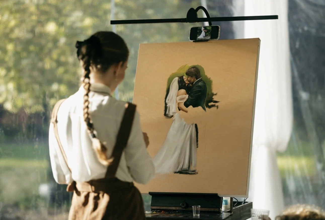 A woman with braided hair painting a portrait of a bride and groom on a canvas outdoors