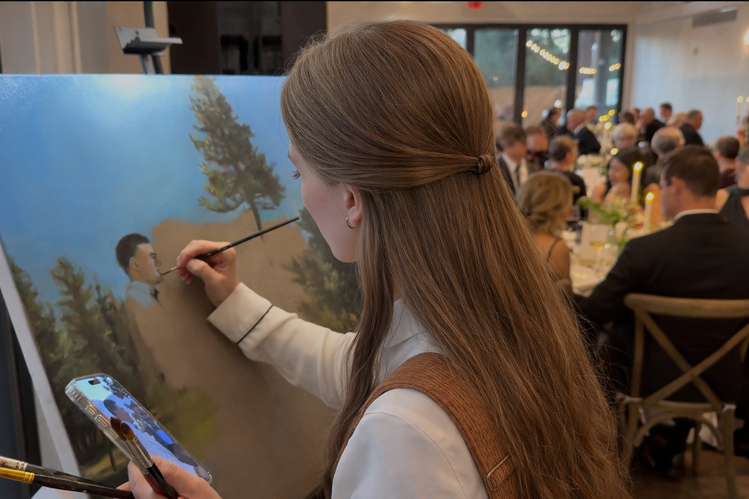 An artist painting a landscape on a canvas at a formal event with round tables and guests in evening attire.