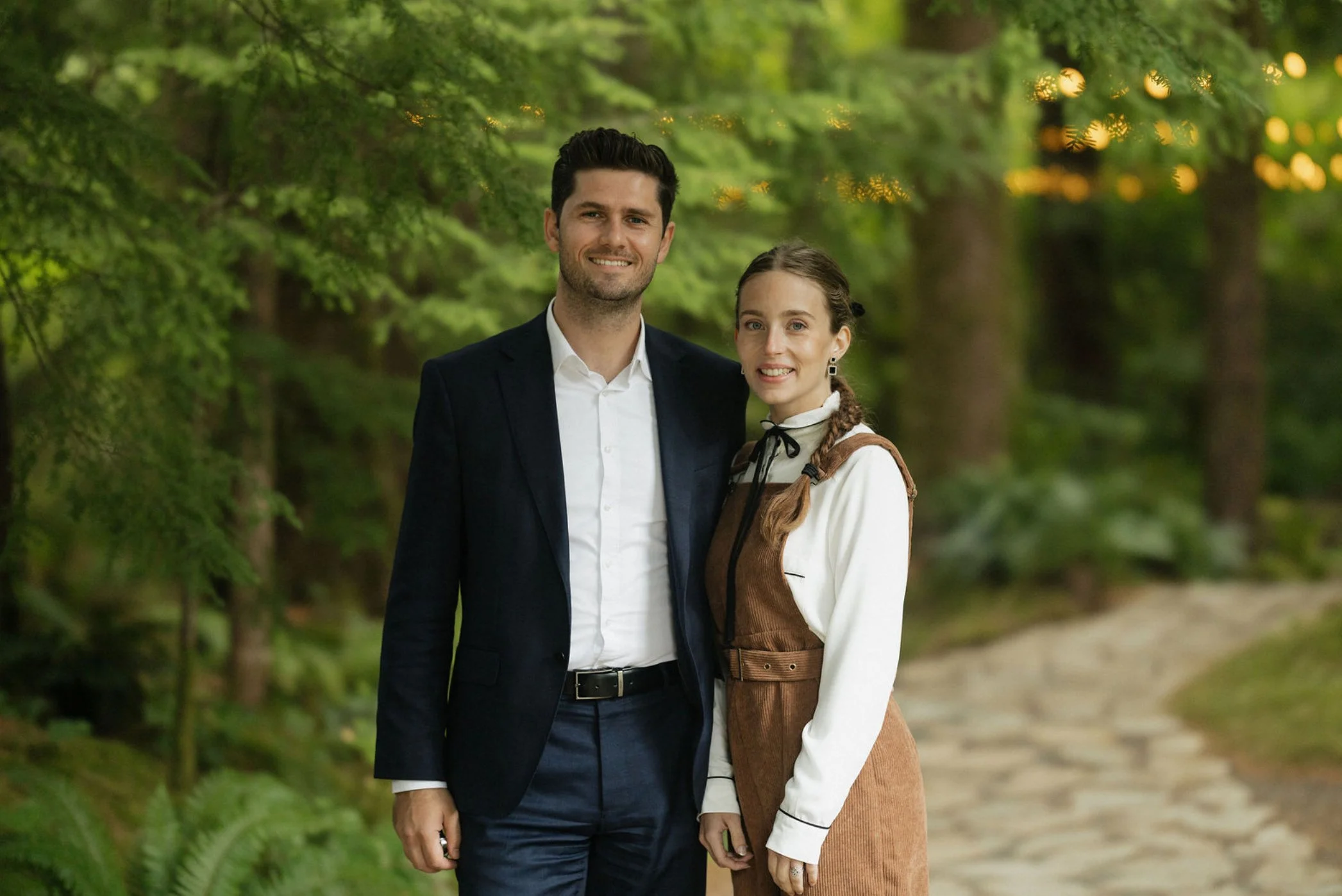 A young man and woman standing together outdoors on a forested path, smiling at the camera.