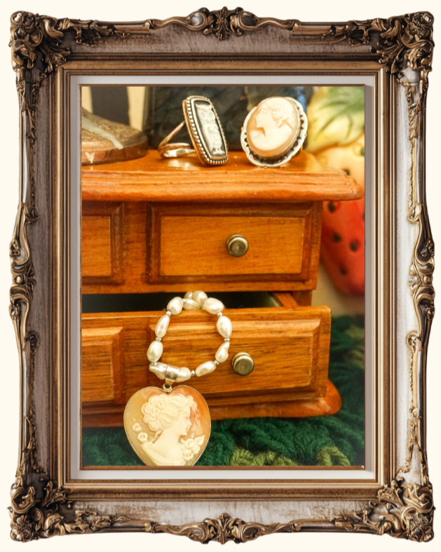 A small wooden jewelry box with two drawers, partially open, resting on a green surface. On top of the box, there are two cameo jewelry pieces with carved profiles and one ring. Hanging from the open drawer, there is a pearl bracelet with a cameo pendant featuring a carved profile of a woman.