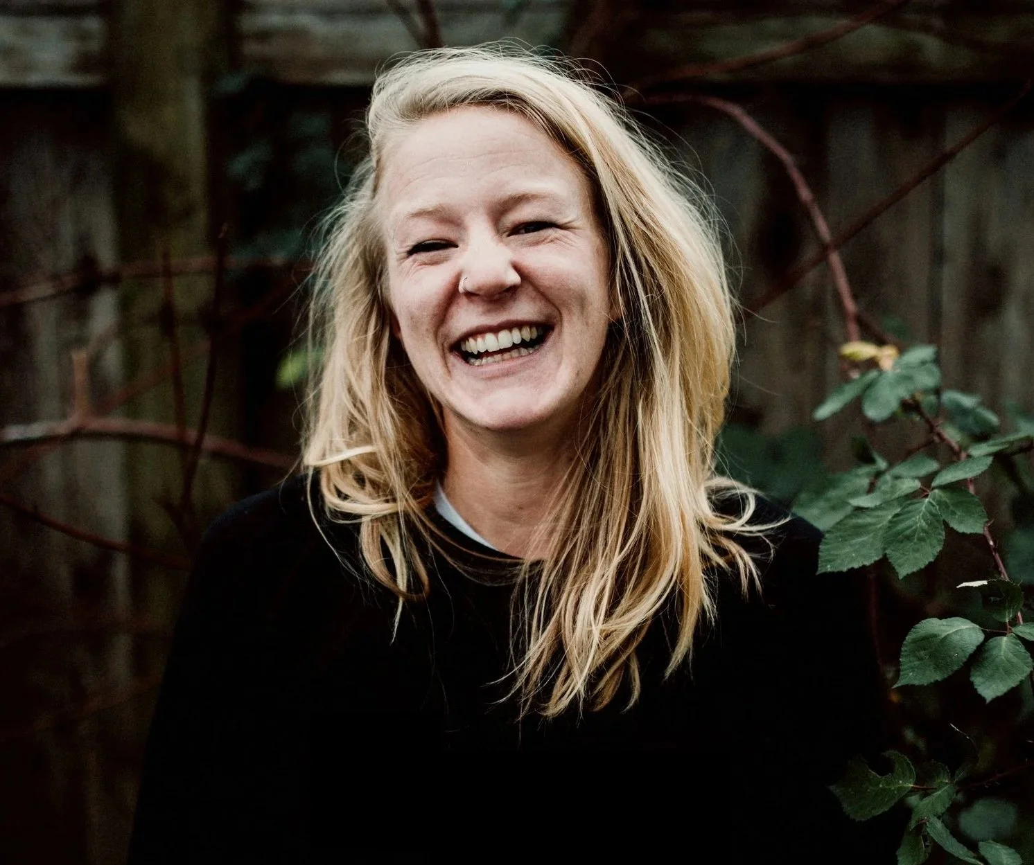 A woman with blonde, wavy hair smiling outdoors in front of a wooden fence and green foliage.