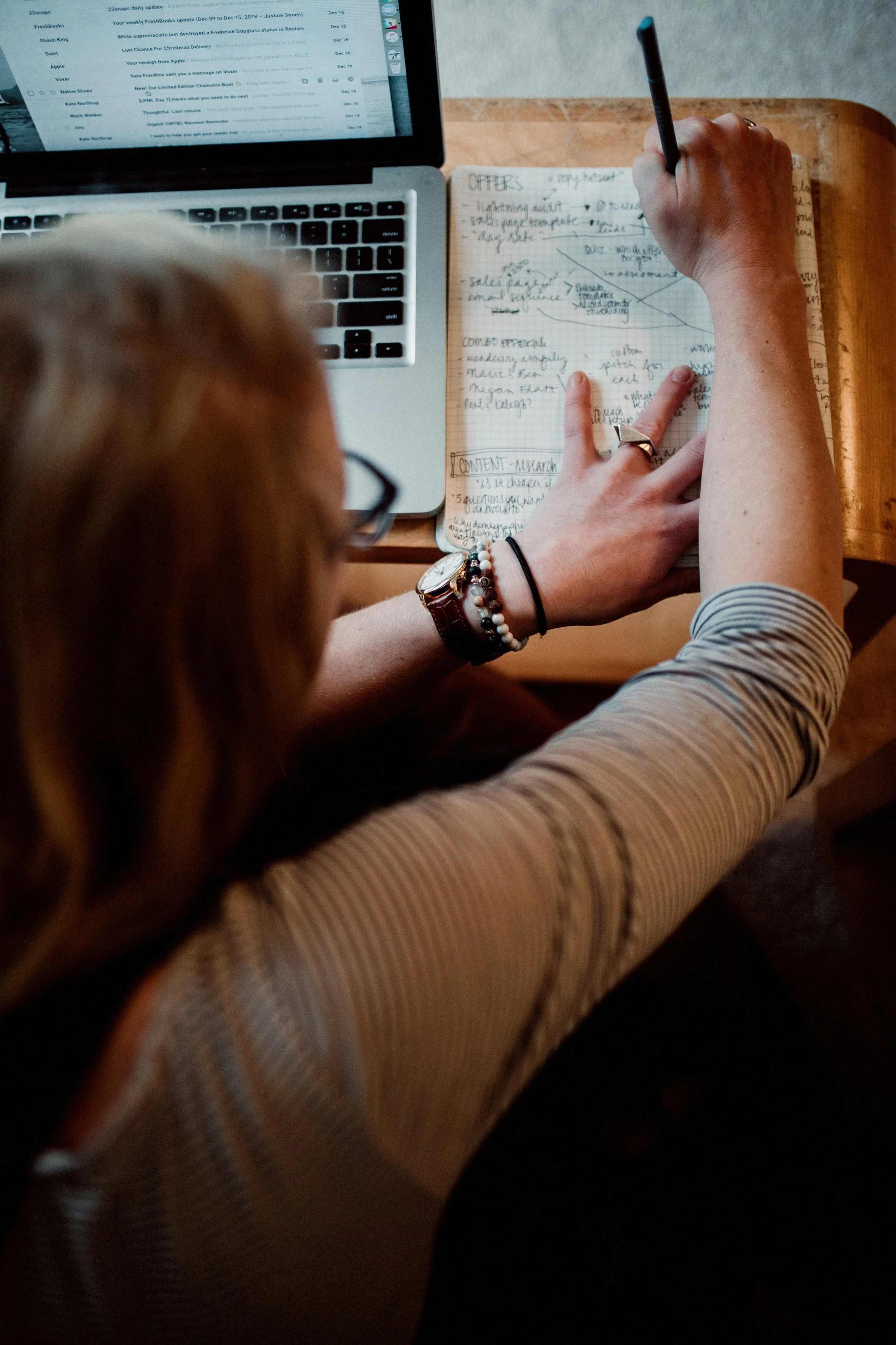 A person with red hair and glasses taking notes in a notebook with a pen, beside a laptop on a wooden table.