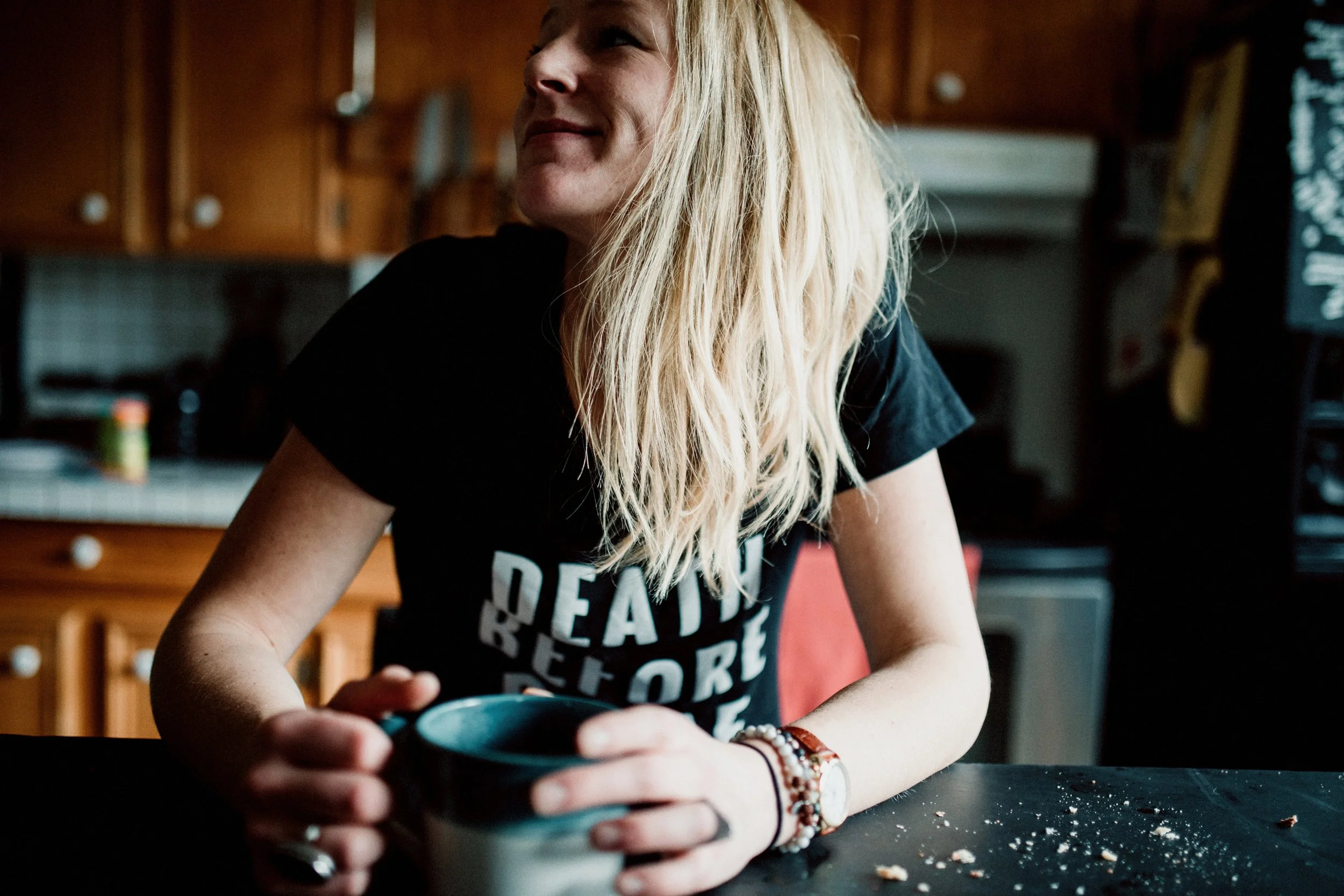 A woman with long blonde hair, wearing a black T-shirt with white text, sitting at a kitchen table holding a mug, with a messy table and a kitchen in the background.