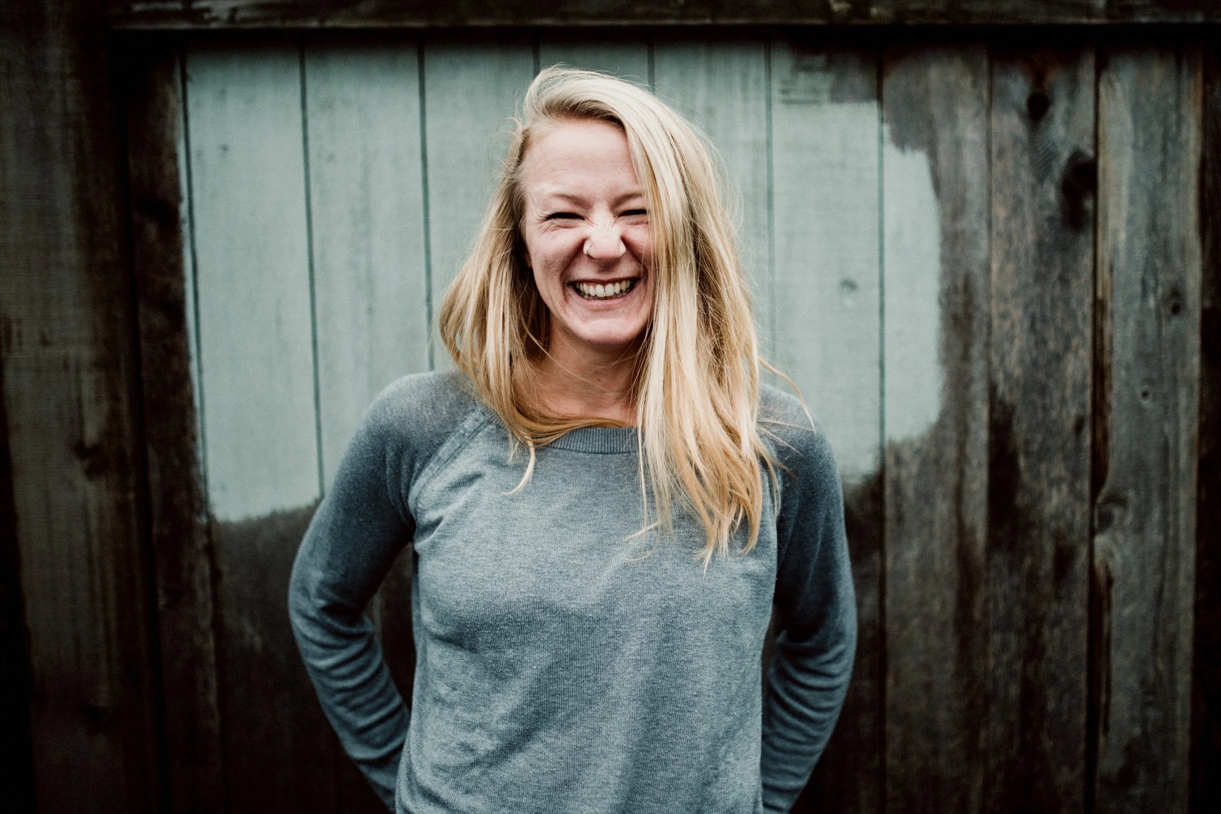 A young woman with blonde hair, smiling and laughing, standing outdoors in front of a wooden fence.