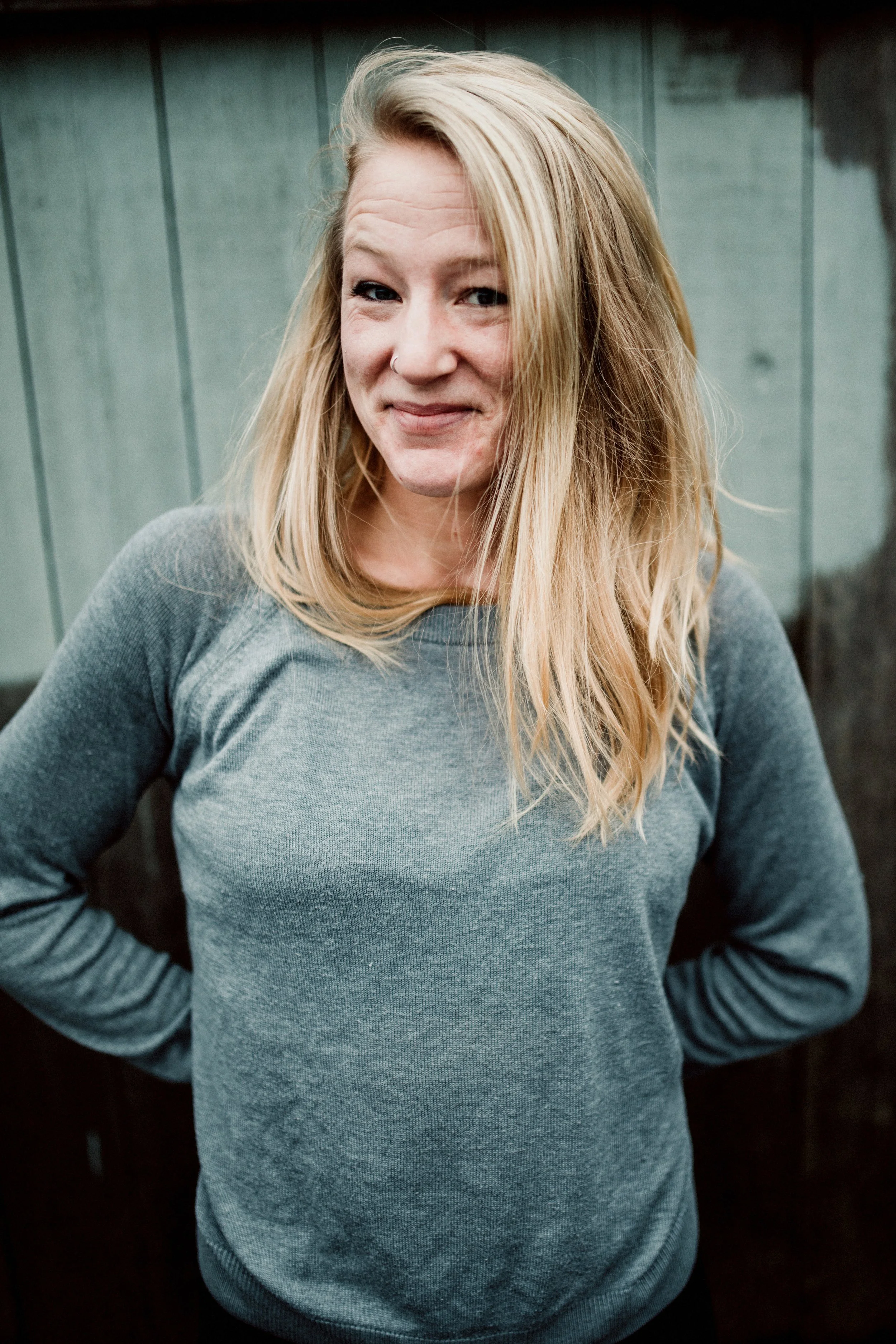 A woman with blonde hair and a nose piercing, wearing a grey long-sleeve shirt, standing outdoors in front of a green wooden wall, smiling slightly with arms behind her back.