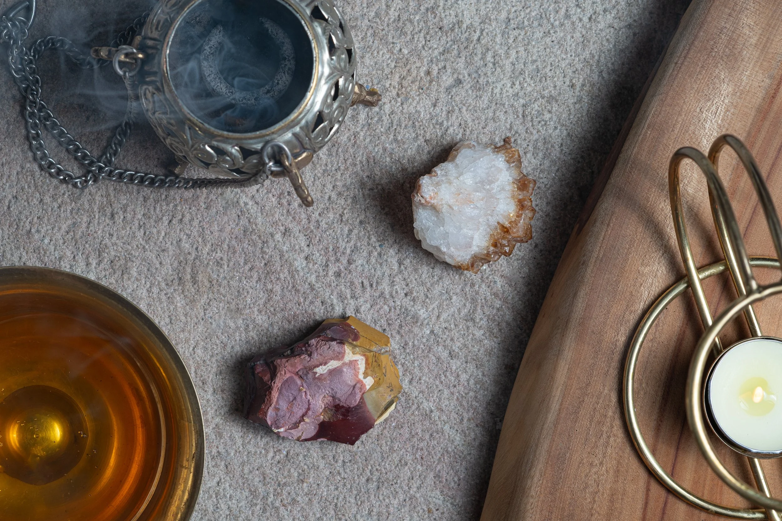 Close-up of a collection of objects on a textured surface, including a small metal lantern, a bowl of amber-colored liquid, two rough stones, a wooden board, and a gold-colored candle holder with a white candle.
