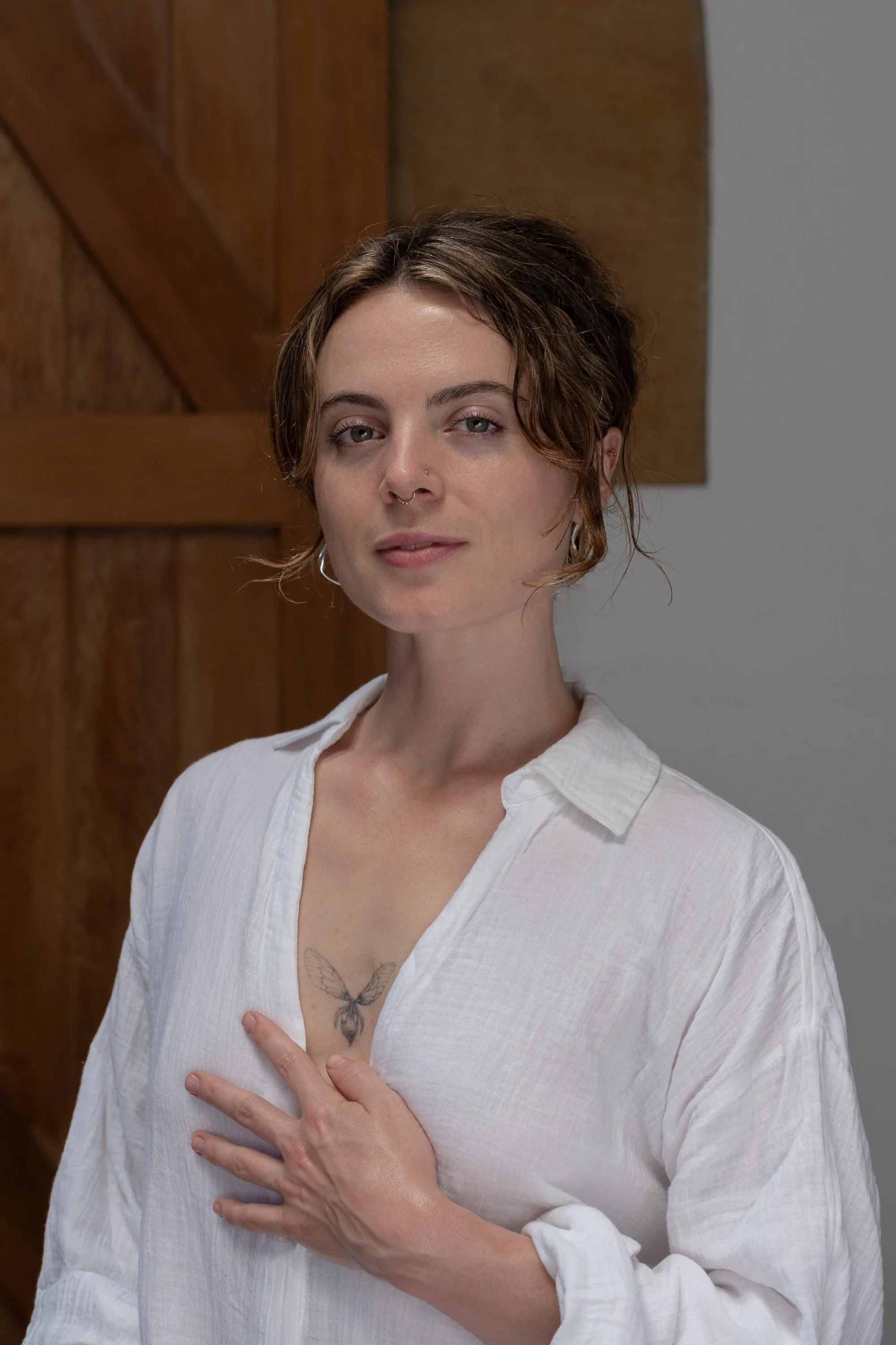 A woman with short, curly brown hair and a septum piercing stands against a wooden wall. She wears a white, loose shirt and has a butterfly tattoo on her chest. Her hand is on her chest as she looks at the camera.