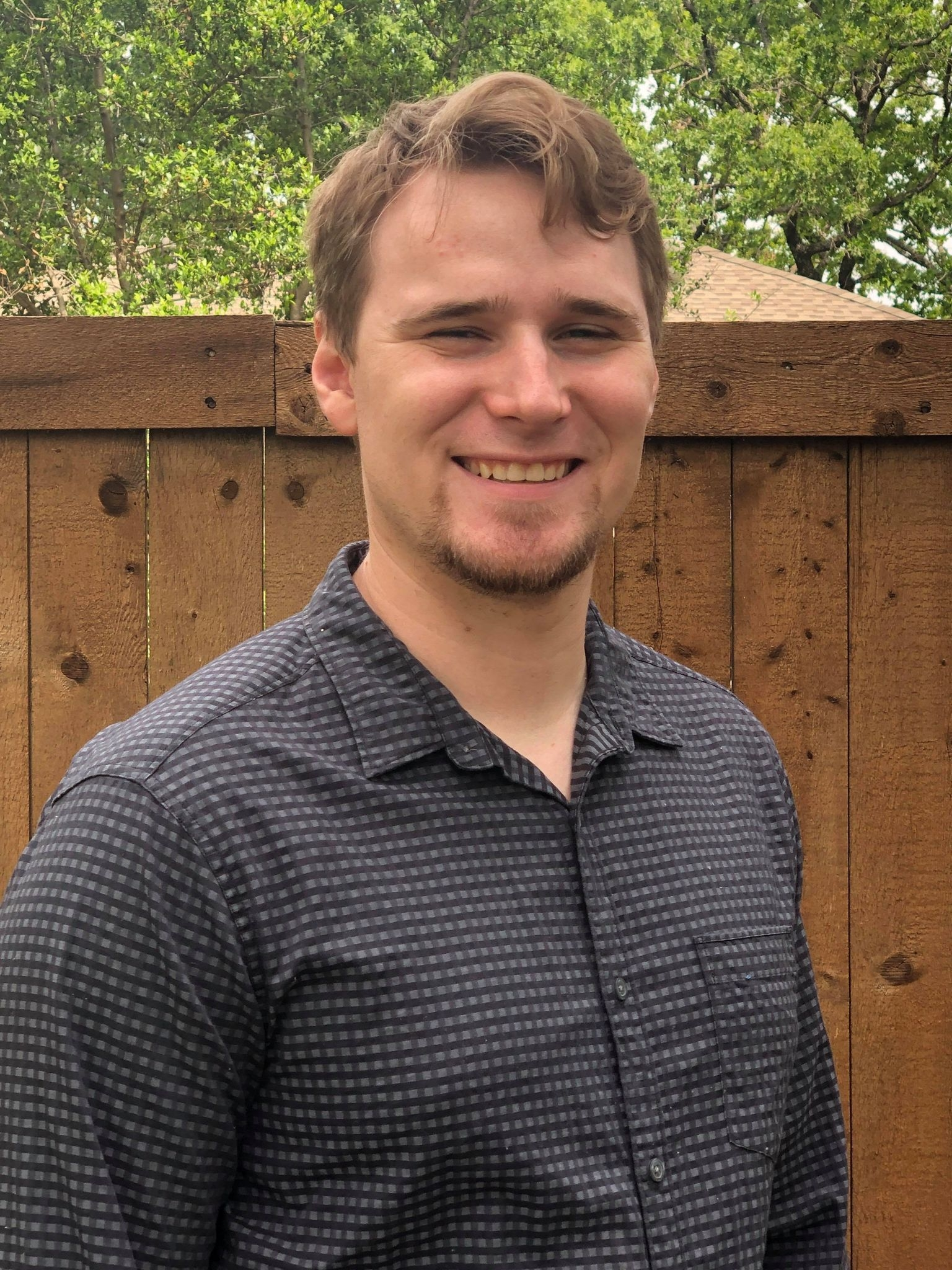 A young man with light skin, short light brown hair, and a goatee, smiling, wearing a checkered dark shirt, standing outdoors in front of a wooden fence with green trees in the background.