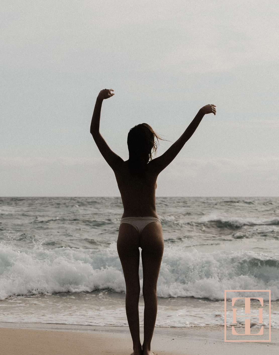 Silhouette of woman standing on beach with arms raised, ocean and cloudy sky in background.