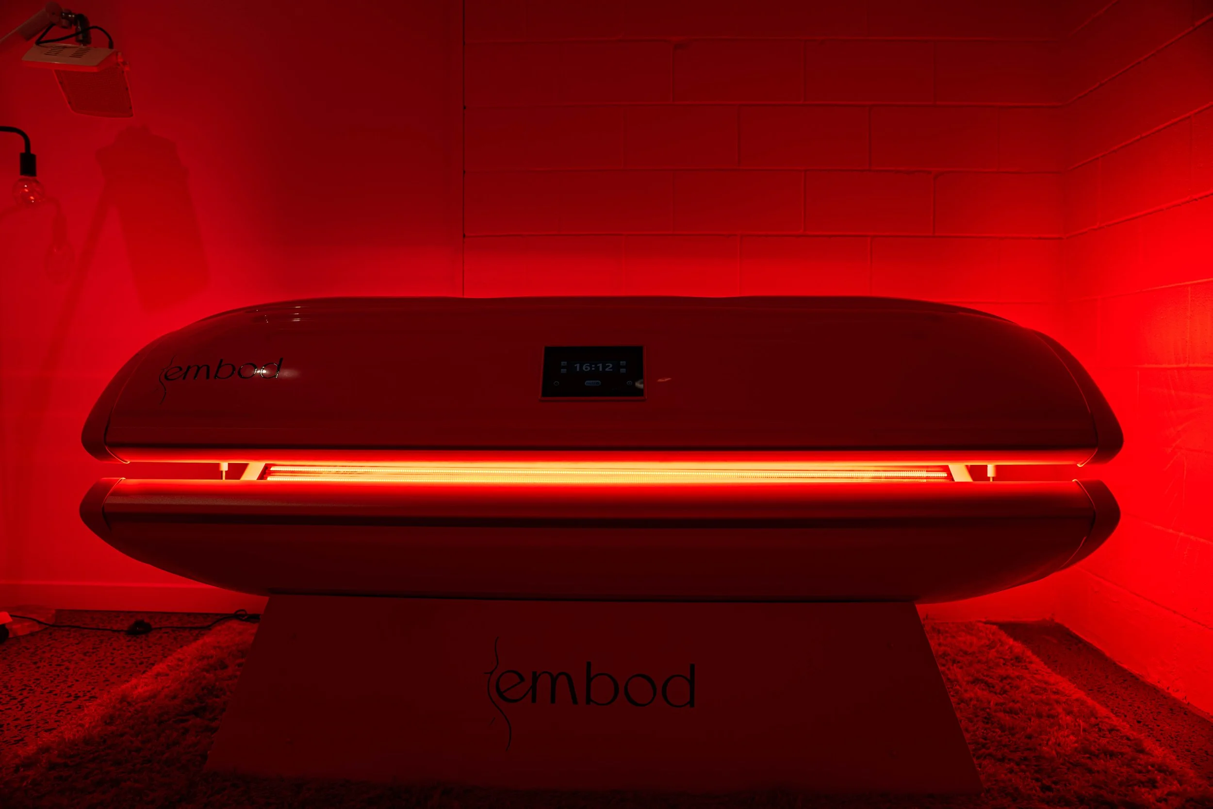 A red light therapy device labeled 'embod' on a white base, emitting a bright red glow in a room with red lighting and brick walls.