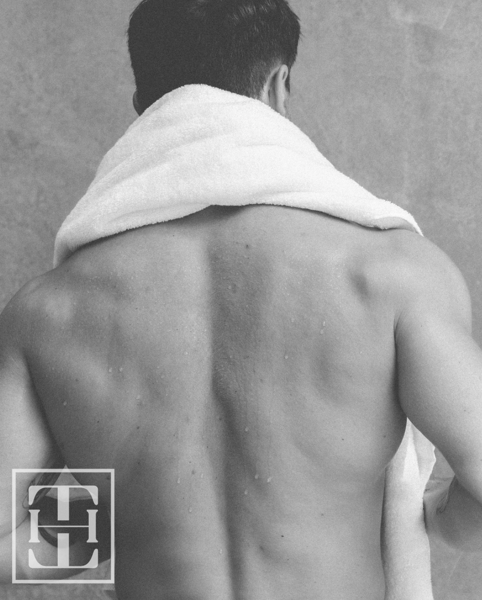 Back view of a shirtless man with a towel draped over his shoulders, standing against a plain background.