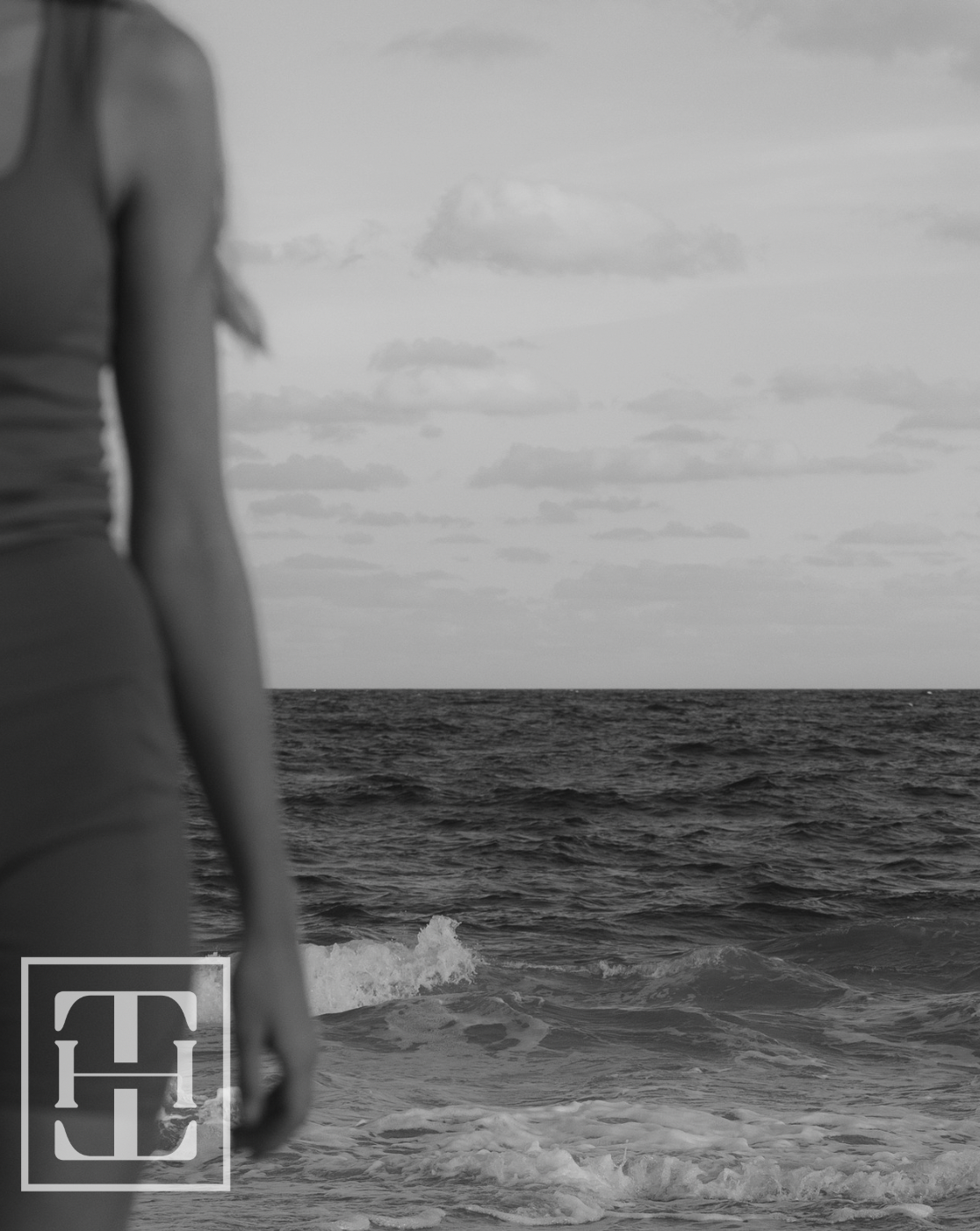 A woman in a swimsuit standing by the ocean, with her back to the camera and topless, at the beach. The photo is in black and white.