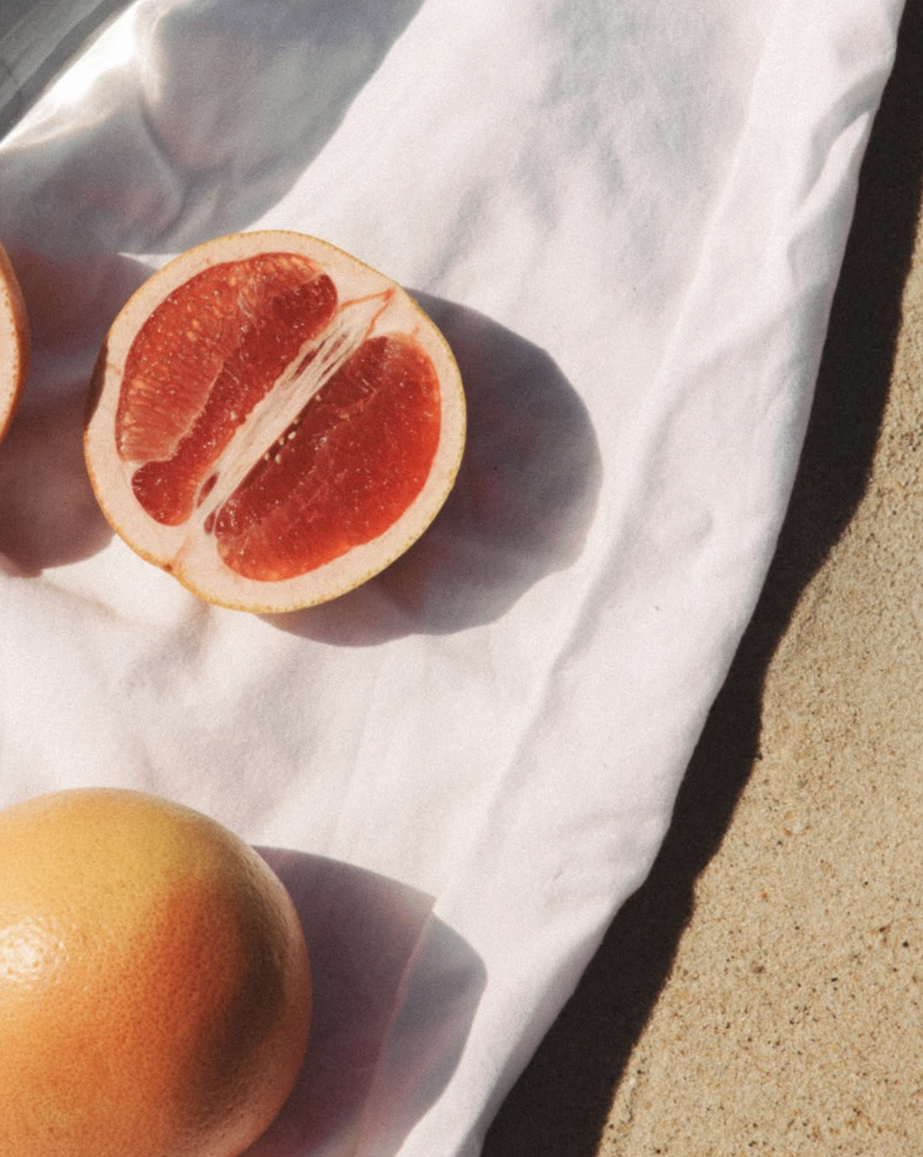 A grapefruit half with pink flesh and a whole grapefruit on white paper on sandy ground.