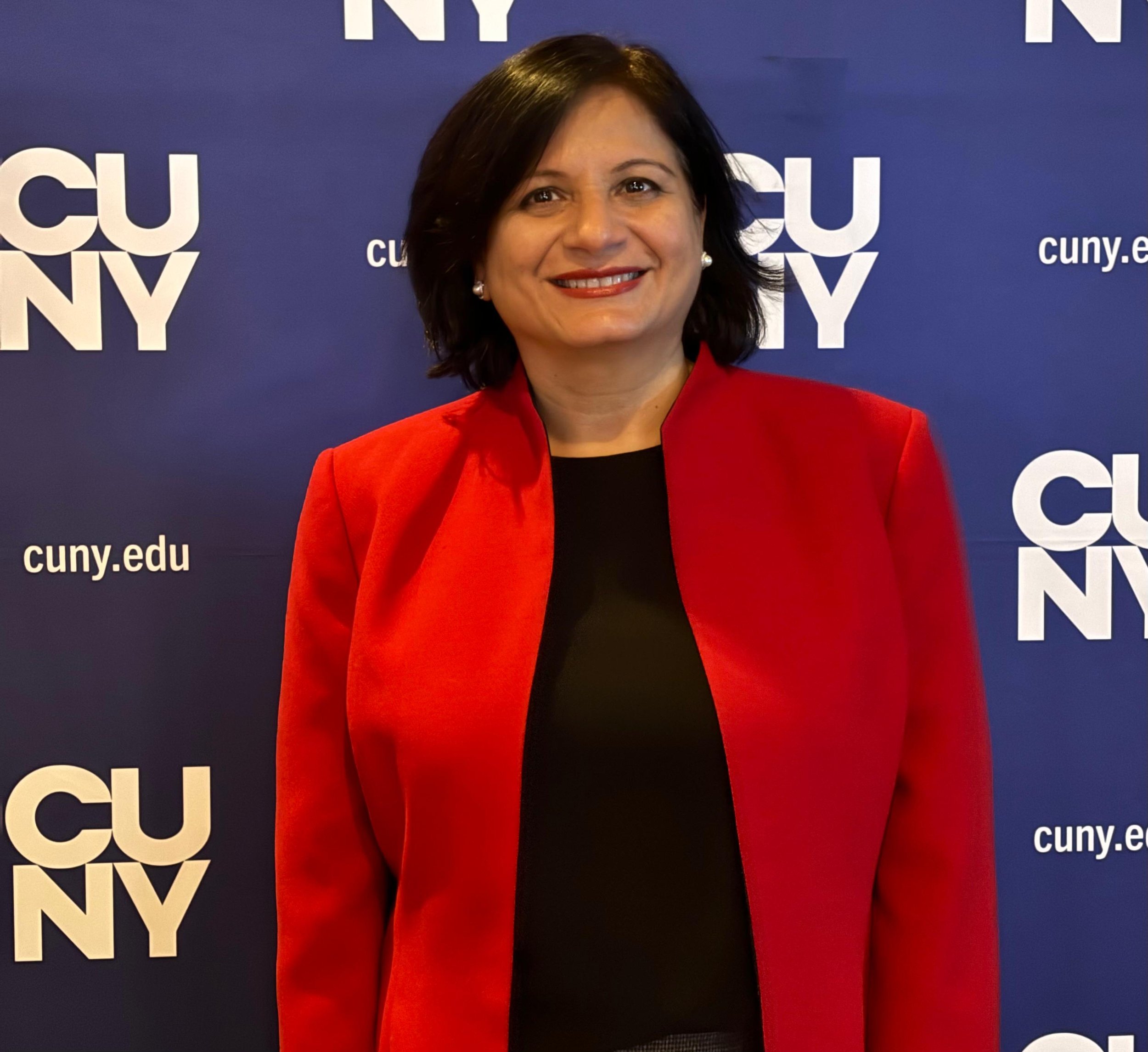 A woman with black hair and pearl earrings smiling, wearing a red blazer over a black top, standing in front of a blue backdrop with white text that says "CUNY" and "cuny.edu."