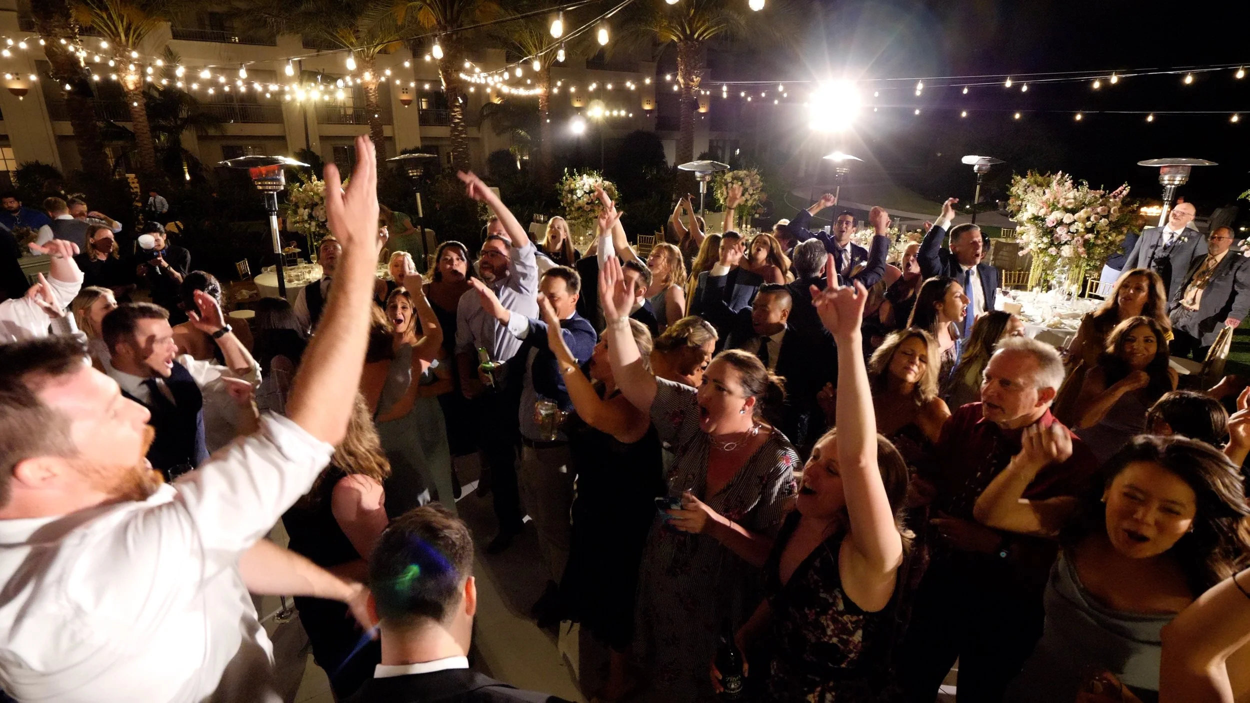 People dancing and celebrating at an outdoor nighttime event with string lights and floral arrangements.